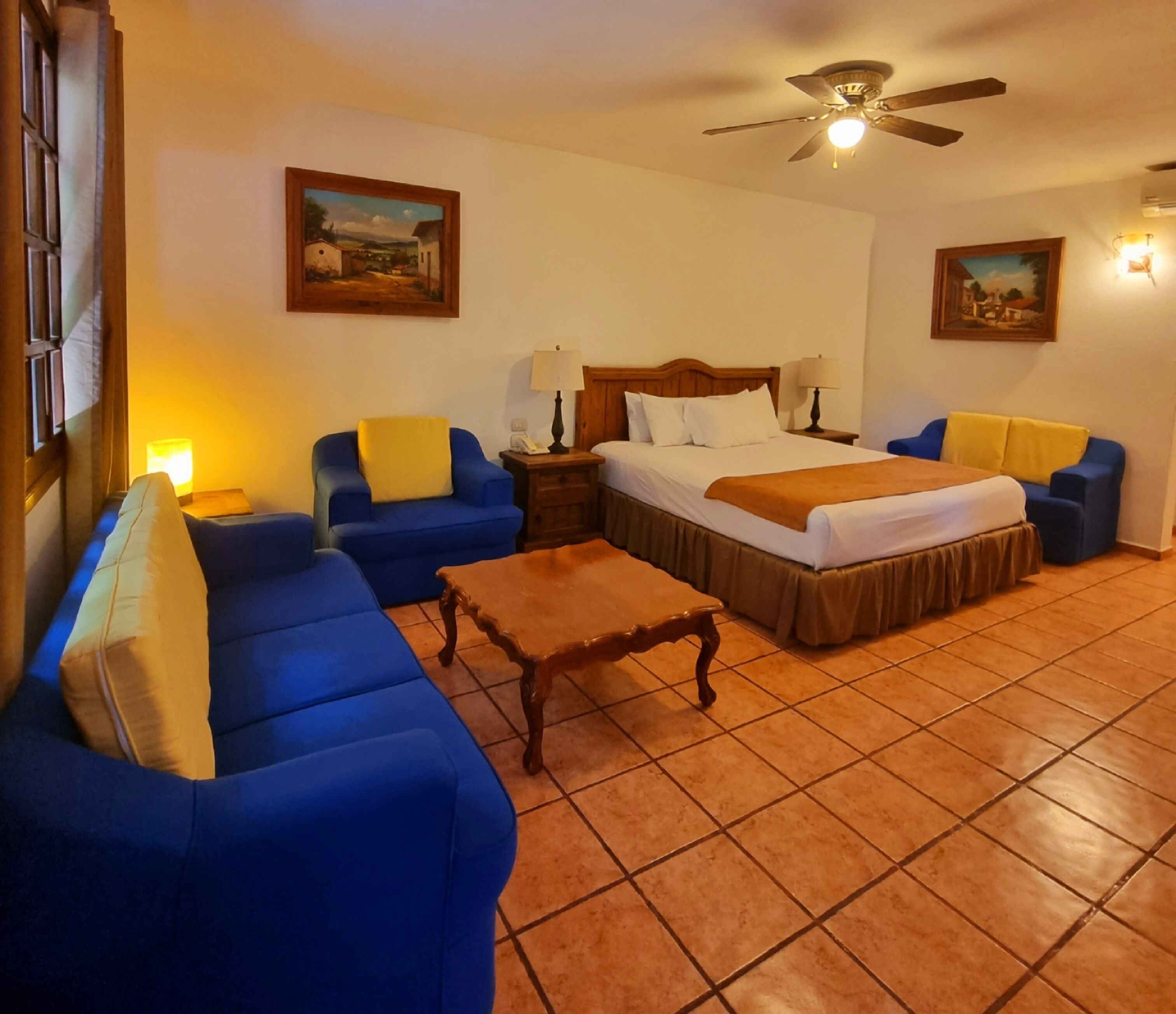 A hotel room with a bed, two blue sofas, a wooden coffee table, and two paintings on the walls.