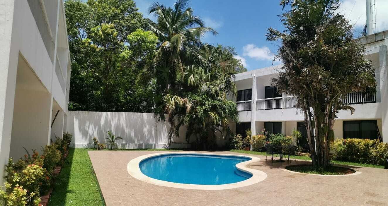 A swimming pool surrounded by greenery and a white building with balconies.