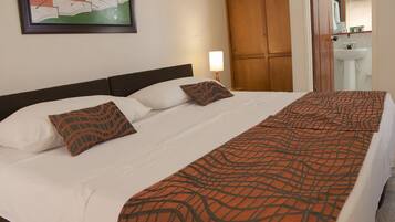 Una cama bien tendida con sábanas blancas y dos almohadas estampadas, un armario de madera y un baño con lavabo y espejo.