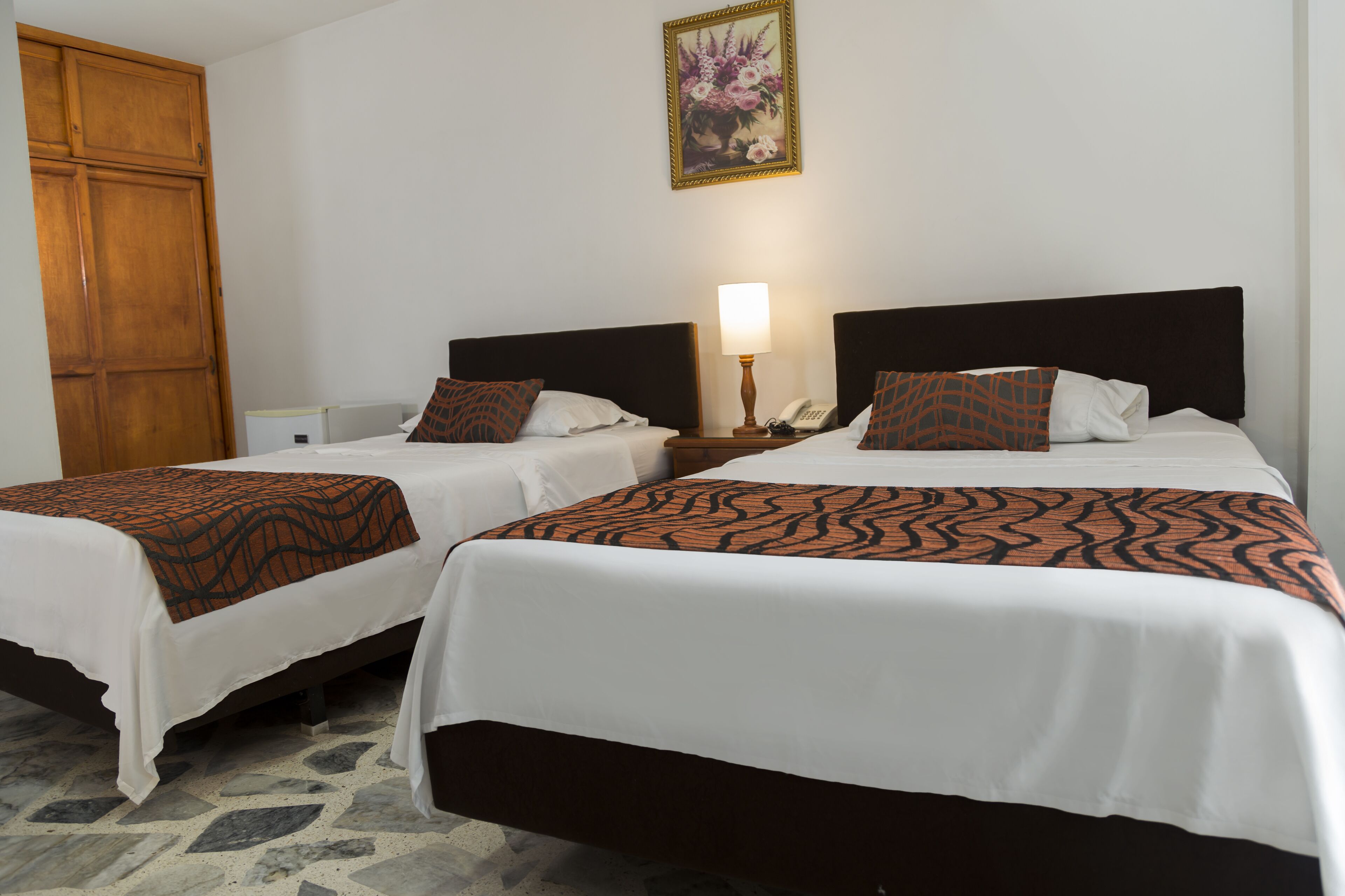 A hotel room with two beds, a wooden wardrobe, a nightstand with a lamp, and a floral painting on the wall.