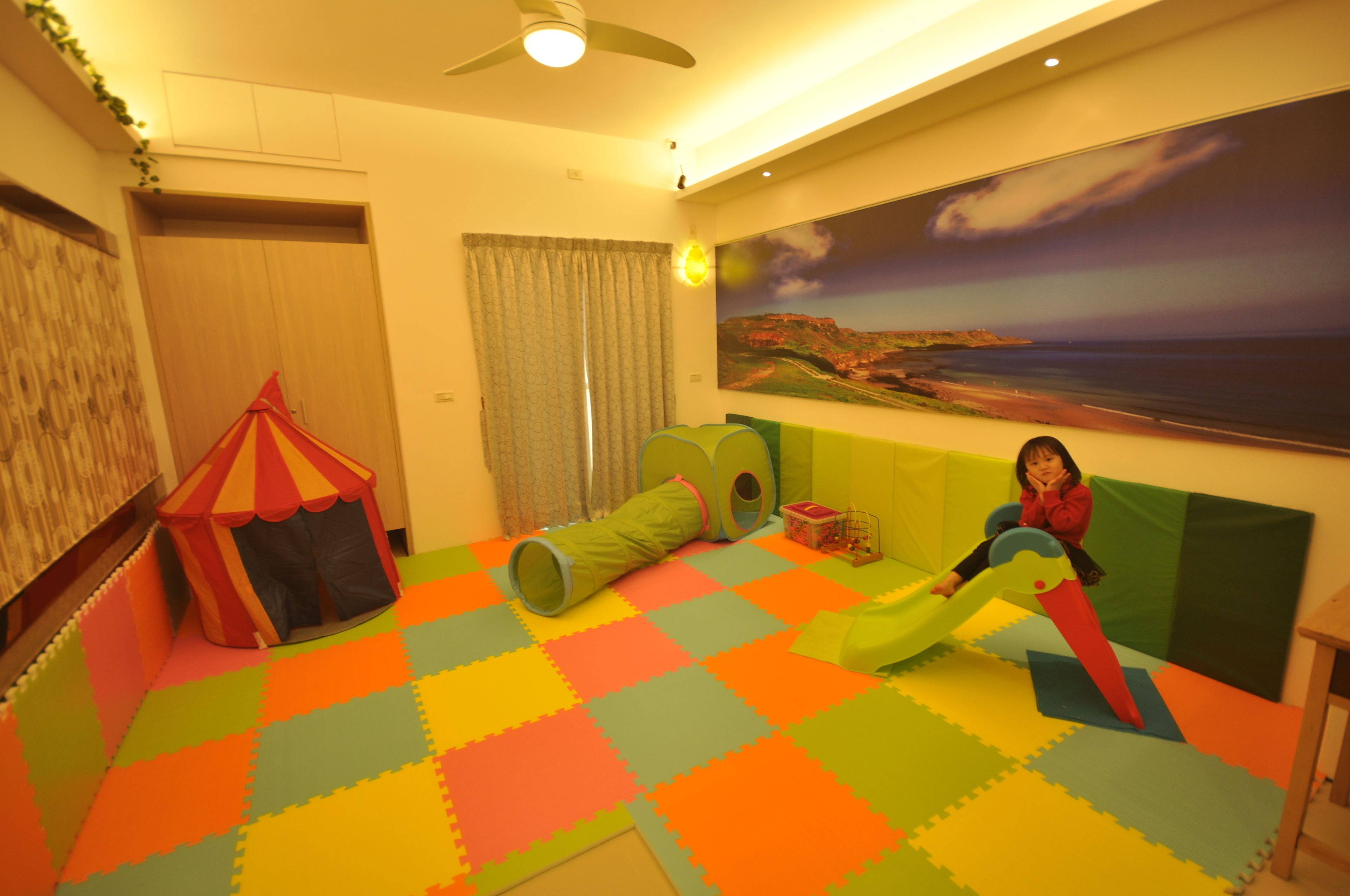 A room with a colorful foam floor, a play tent, a green tunnel, and a large landscape painting on the wall.