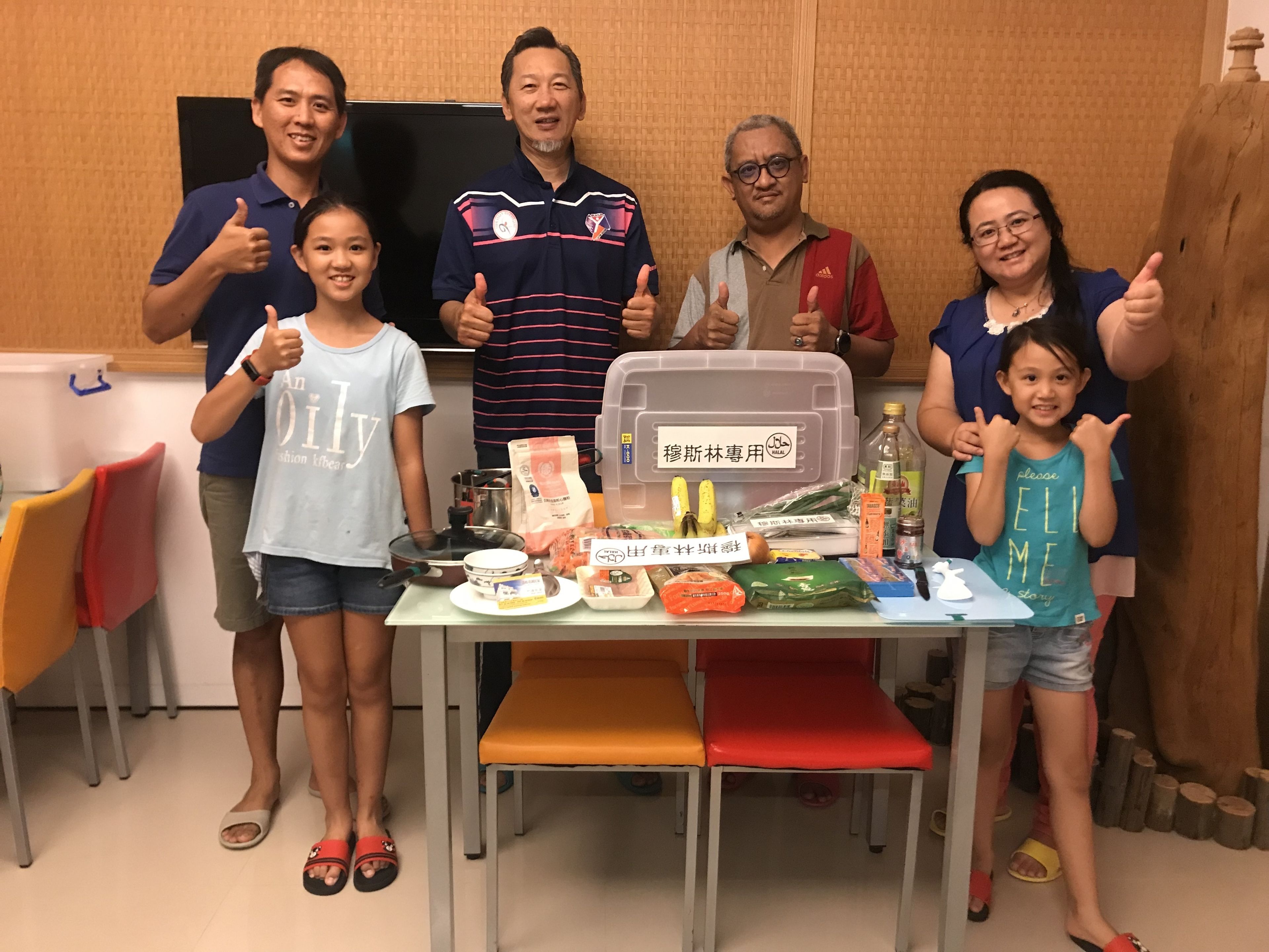 Un grupo de personas de pie alrededor de una mesa con diversos alimentos y un cartel en una habitación con paredes de madera y una pizarra.