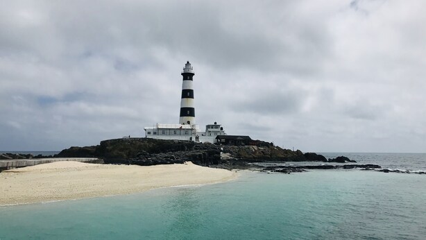 Un faro en una isla rocosa con una playa de arena en primer plano y un cielo nublado.