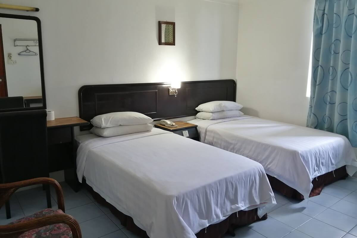 A hotel room with two single beds, a desk with a chair, a bedside table, a mirror, and a window with curtains.