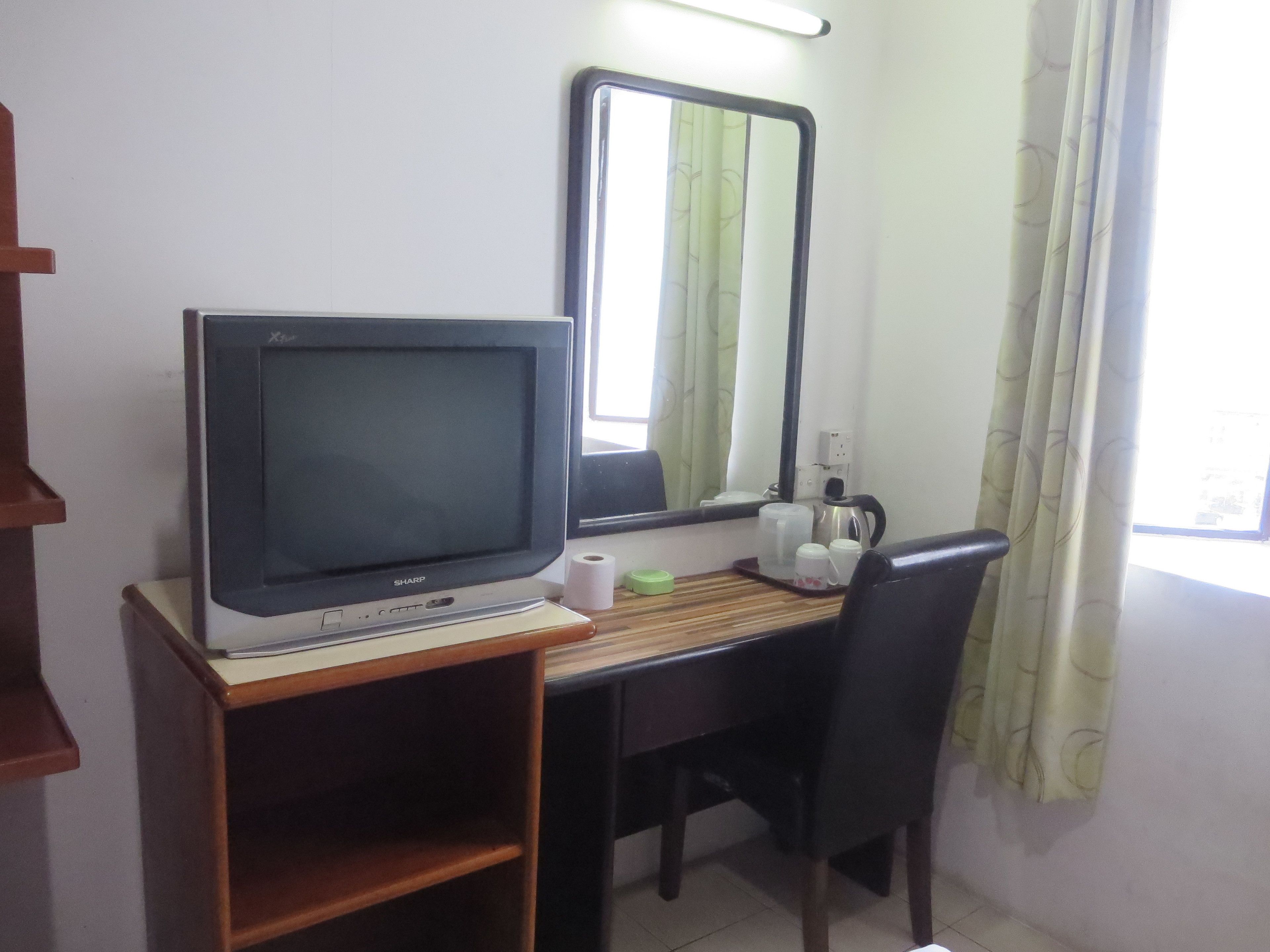 A room with a desk, a computer monitor, a chair, a mirror, a window with curtains, and a shelf with a roll of toilet paper.