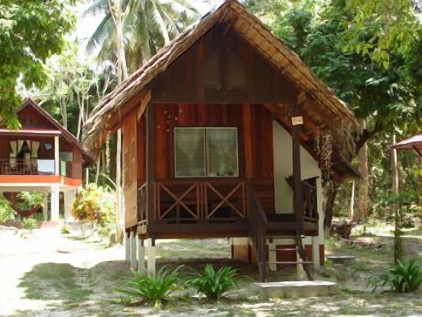 A wooden bungalow with a thatched roof, surrounded by lush greenery and palm trees.