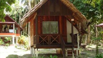 A wooden bungalow with a thatched roof, surrounded by lush greenery and palm trees.