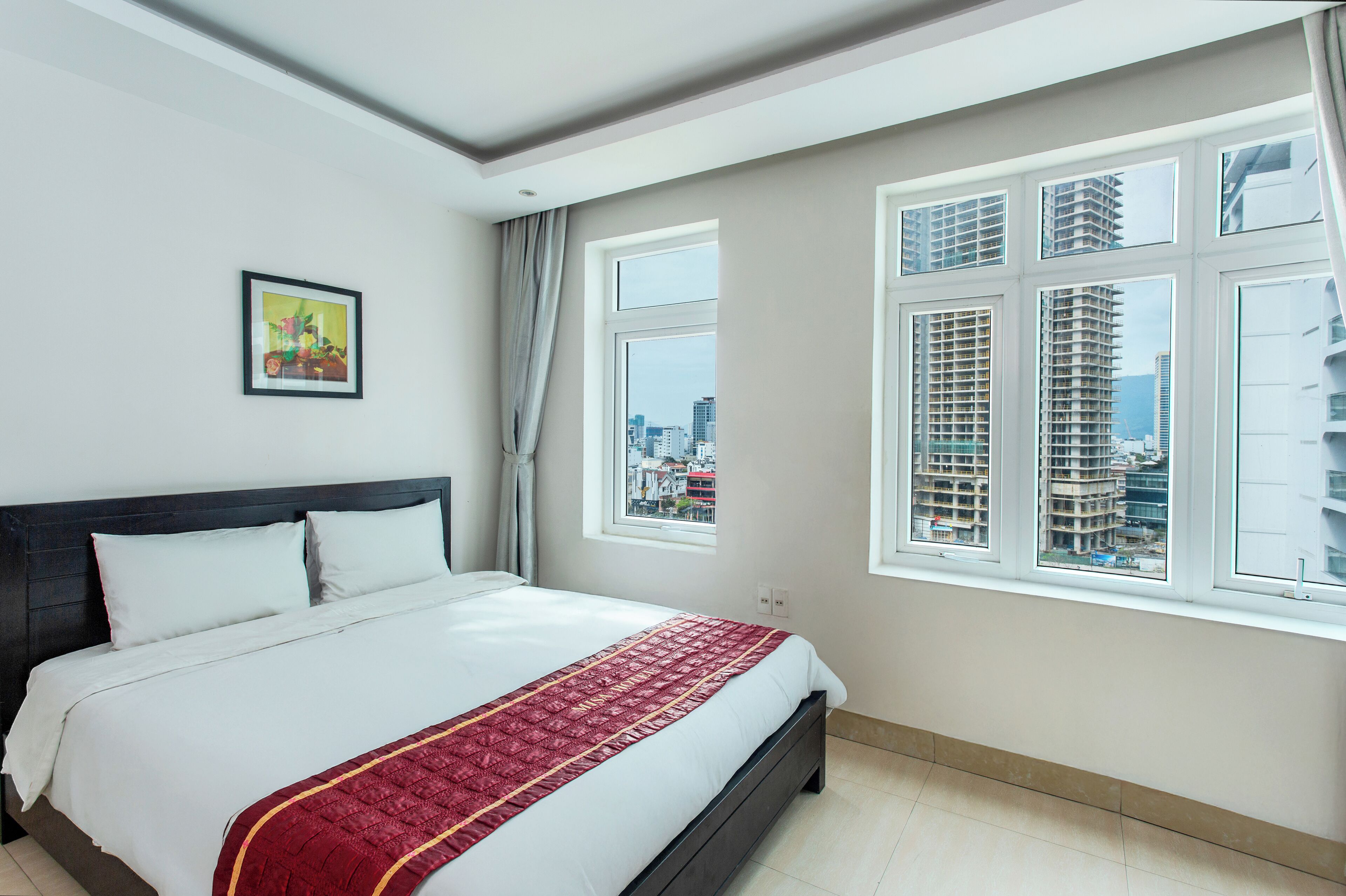 Une chambre d’hôtel avec un grand lit, une vue sur la ville par les fenêtres et un tableau encadré au mur.