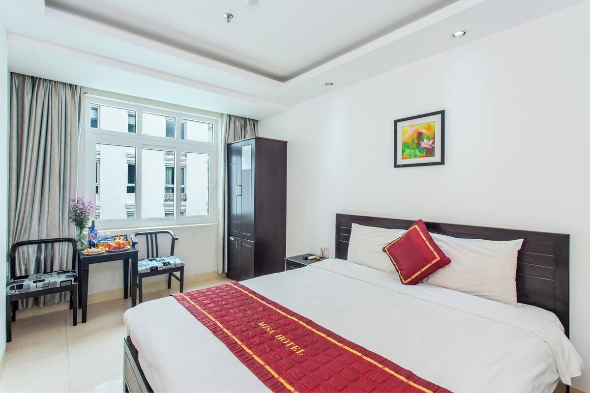 A hotel room with a large bed, a desk with a chair, a wardrobe, and a window with curtains.