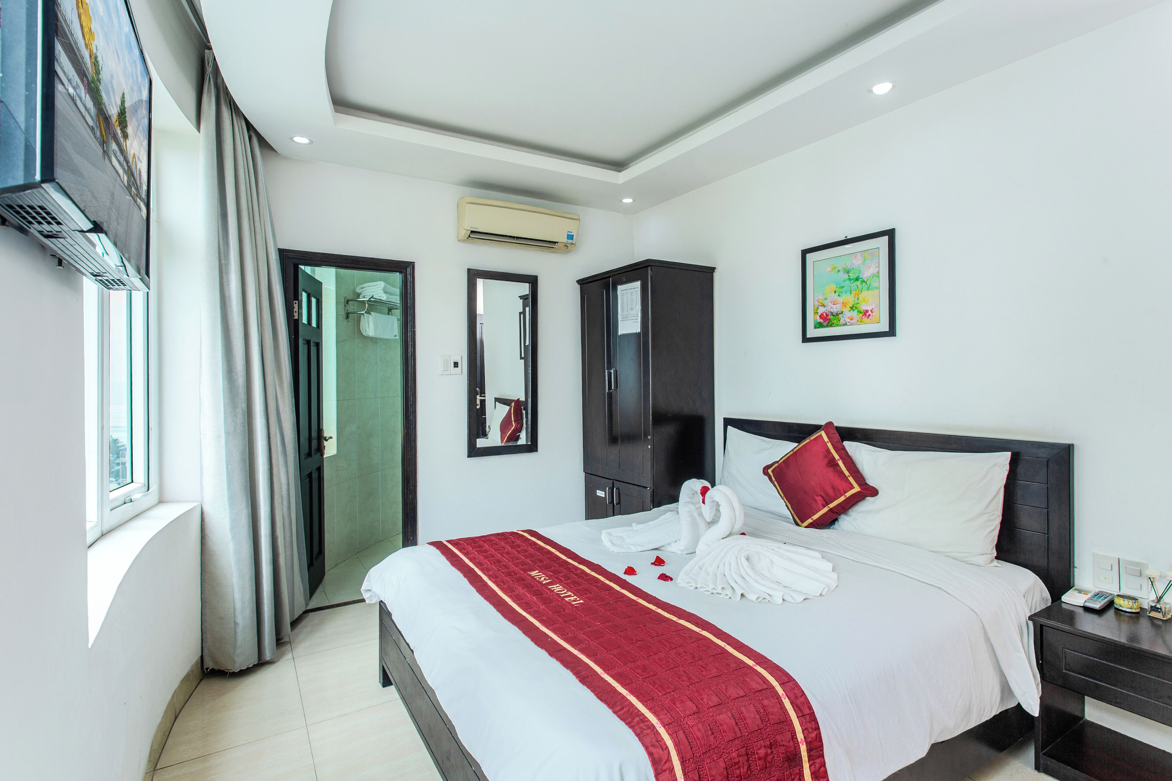 A hotel room with a bed, a wardrobe, a nightstand, a mirror, an air conditioning unit, and a window with curtains.