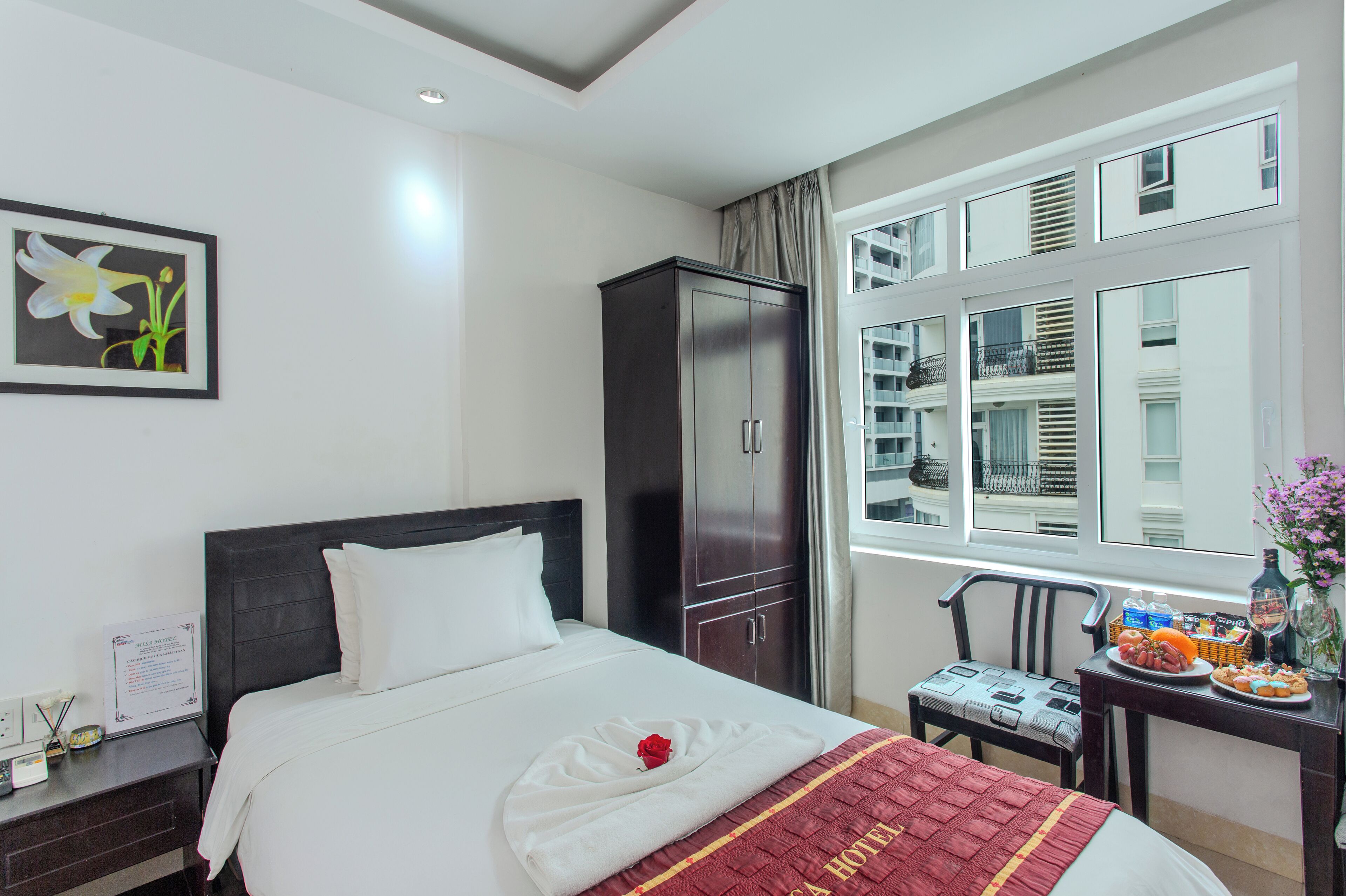 A hotel room with a bed, a wardrobe, a desk with a vase, and a window with a view of buildings.