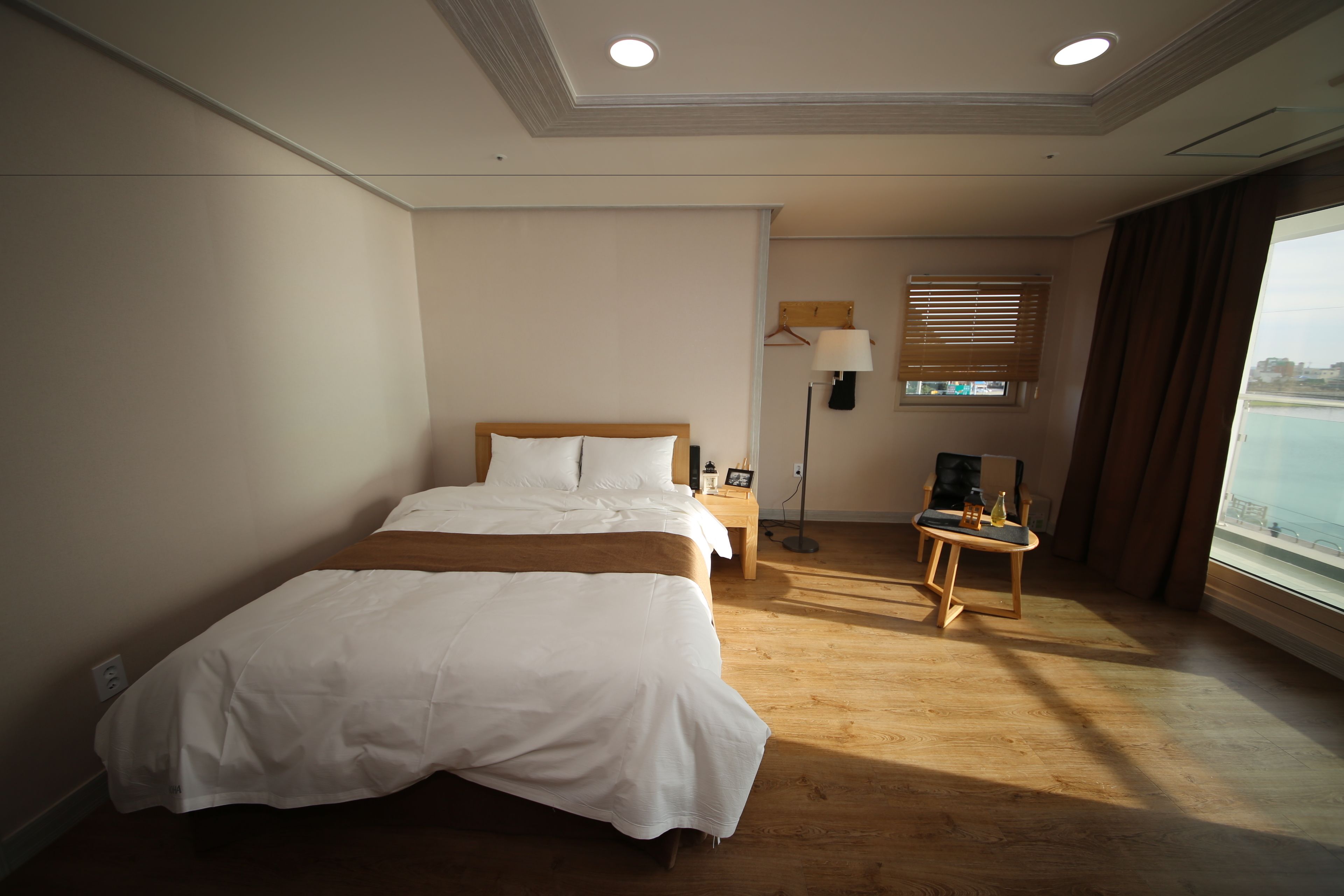 A hotel room with a large bed, a bedside table, a lamp, a window with blinds, and a view of the sea.