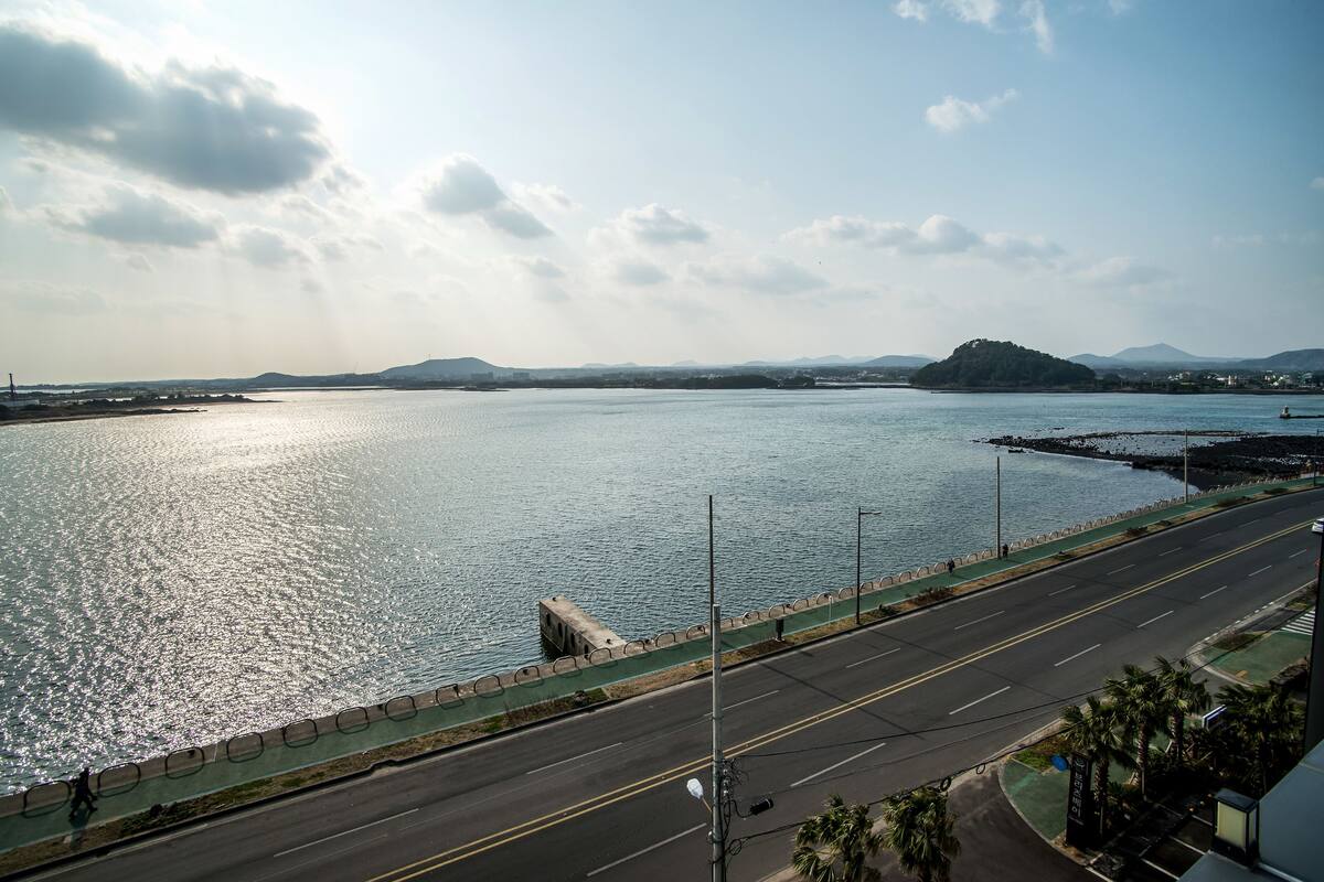 A coastal road with a pedestrian path alongside a body of water, a bridge, and distant hills.