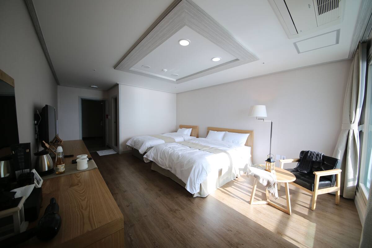 A modern hotel room with a wooden floor, a large bed with white linens, a desk with a laptop, a chair, and a small table with a lamp.