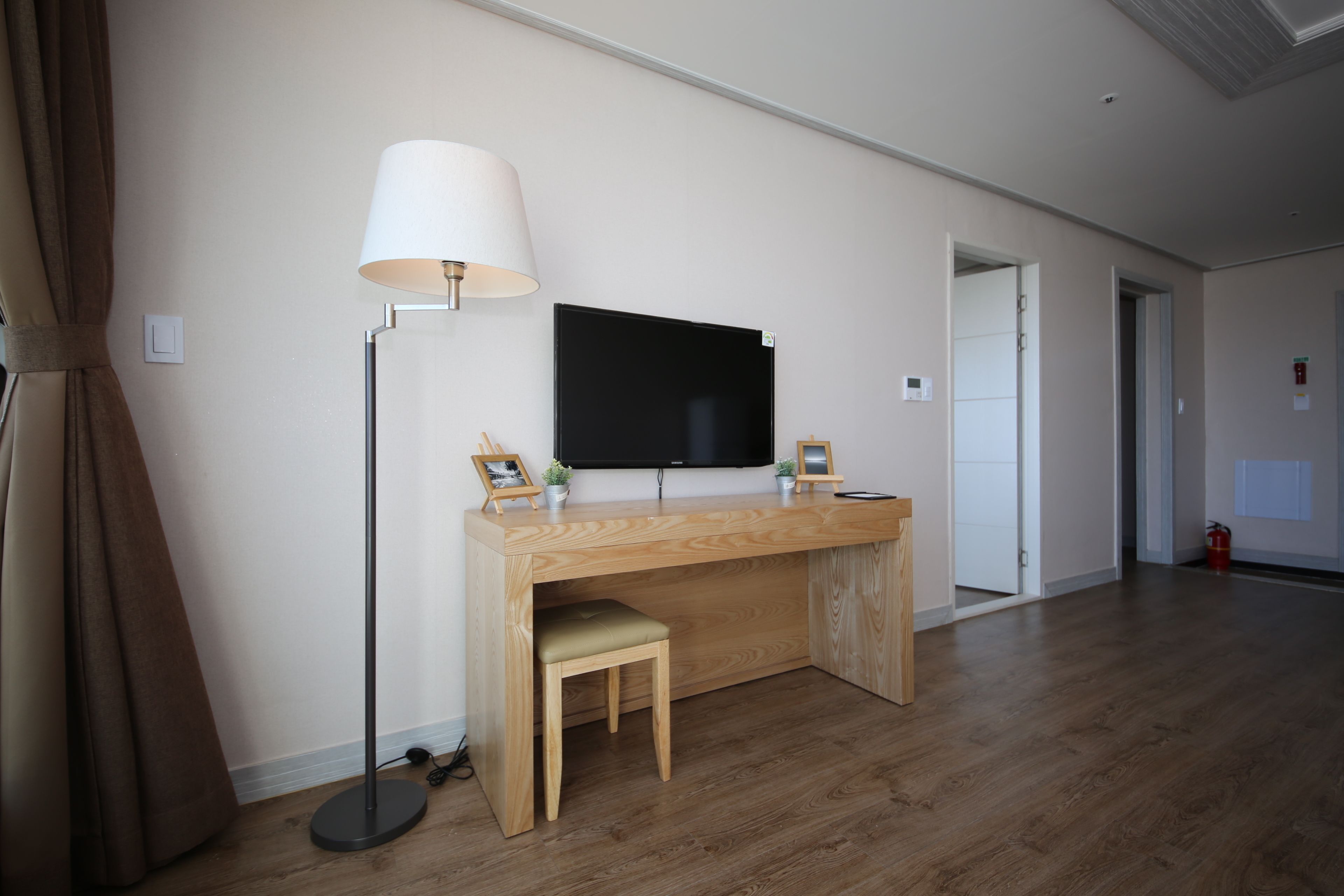 Een kamer met een houten bureau, een zwarte flatscreen-tv, een vloerlamp en een klein krukje.