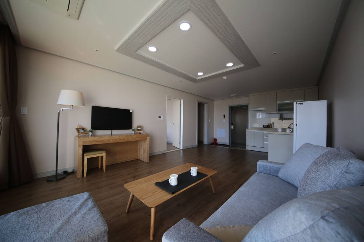 A modern living room with a wooden coffee table, grey sofas, a flat-screen TV, and a kitchen in the background.