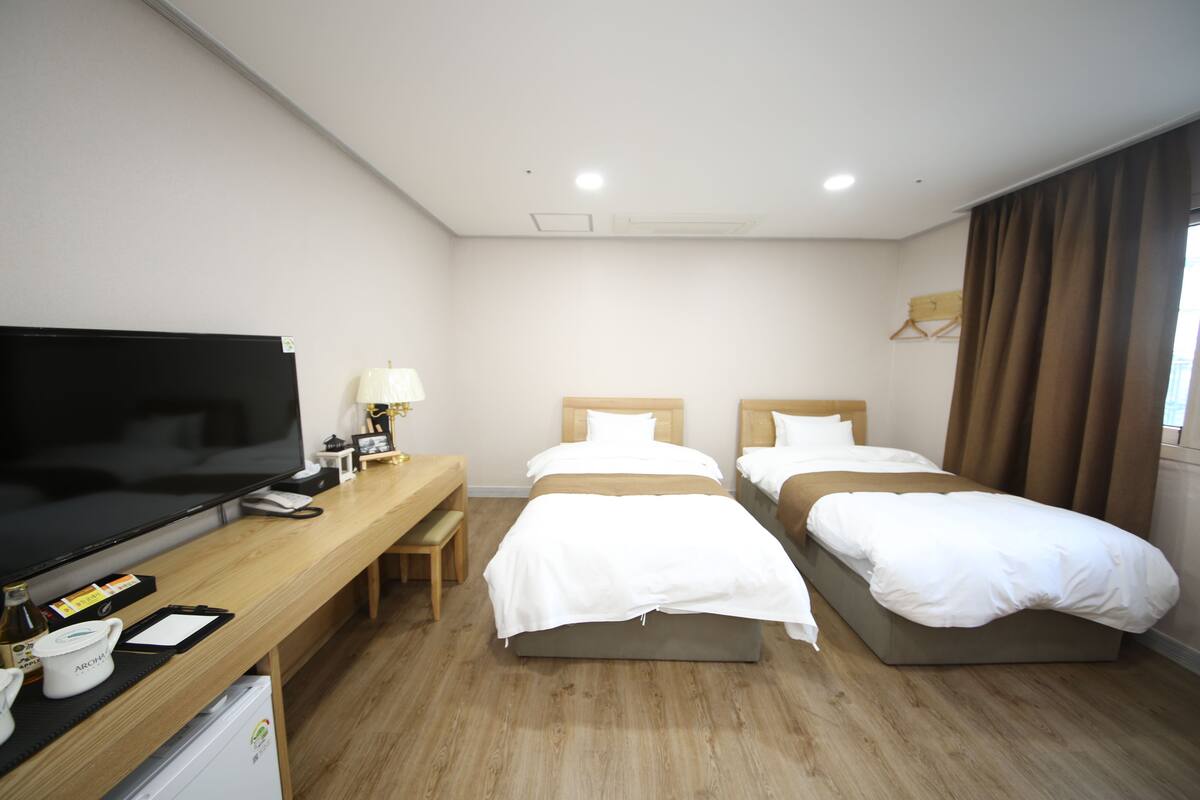 A hotel room with two single beds, a large flat-screen TV, a wooden desk with a chair, and a small refrigerator.