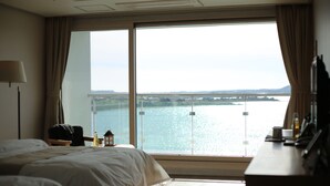 Habitación de hotel con un ventanal grande que ofrece vistas a un cuerpo de agua y un puente. Cuenta con una cama con ropa blanca, un escritorio con lámpara y una silla.