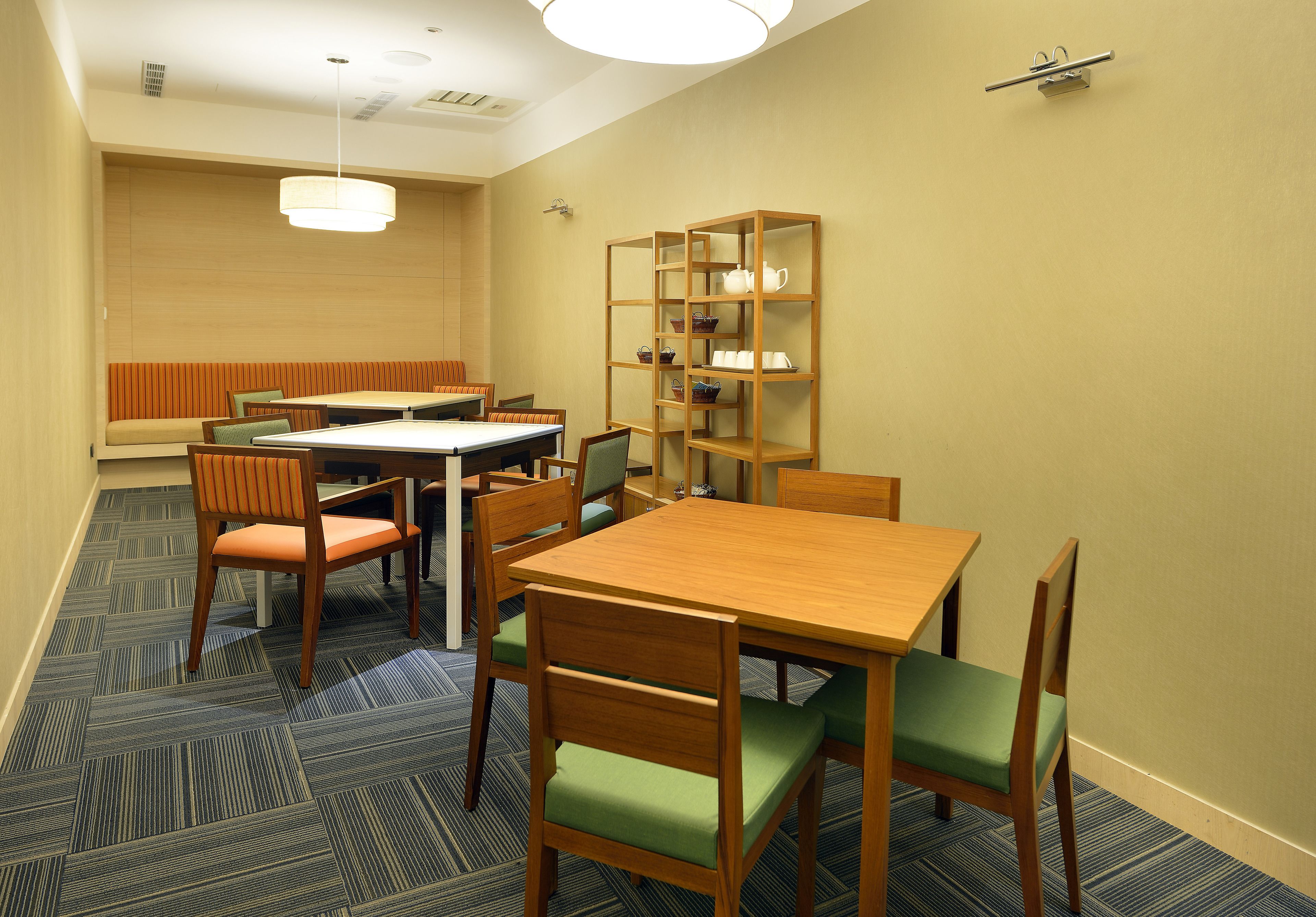 A hotel room with a dining area featuring a wooden table and chairs, a bookshelf, and a patterned carpet.