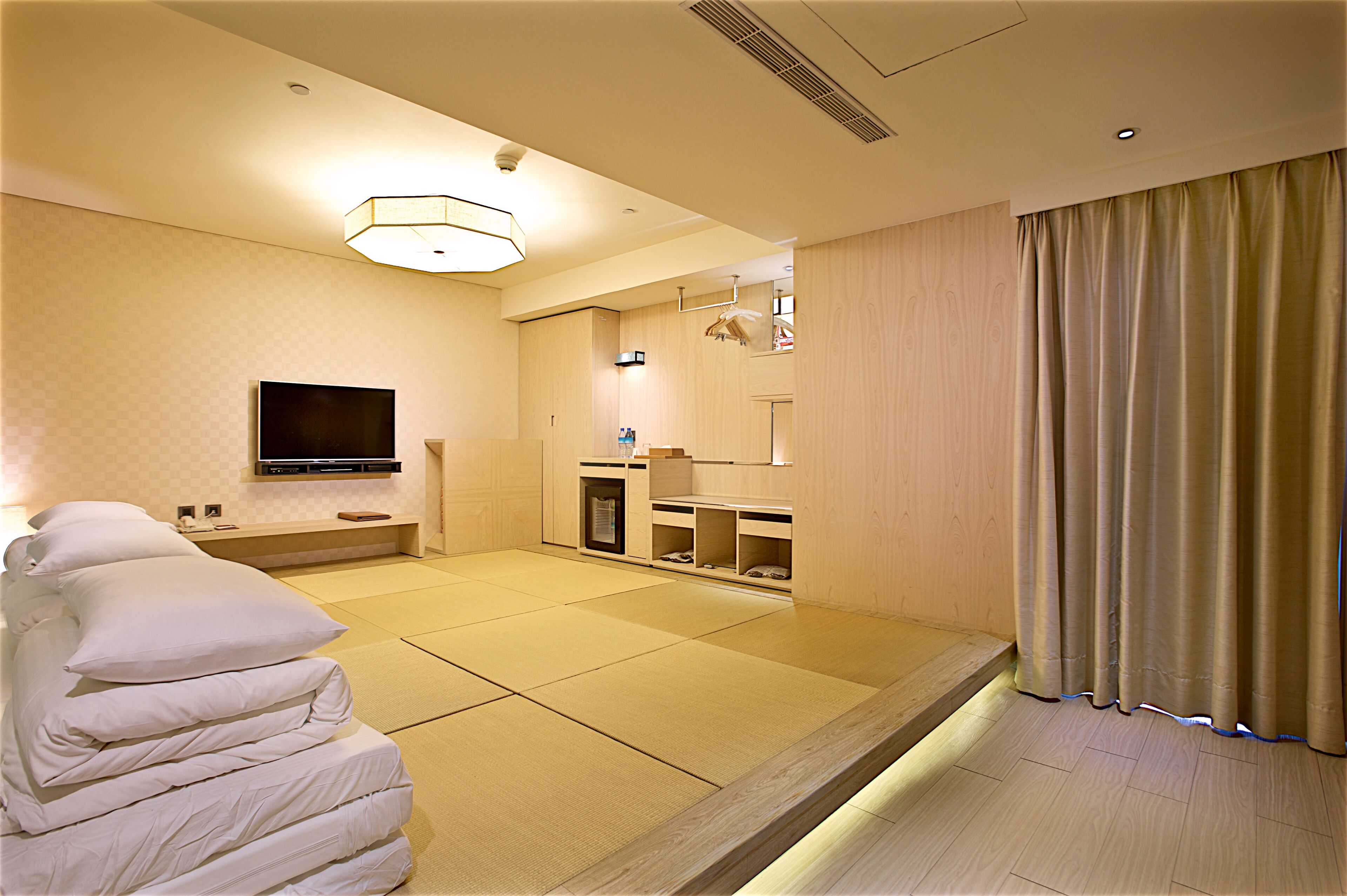 Habitación de hotel con suelo de tatami, televisor montado en la pared, cama con almohadas, una mesita y una ventana con cortinas.