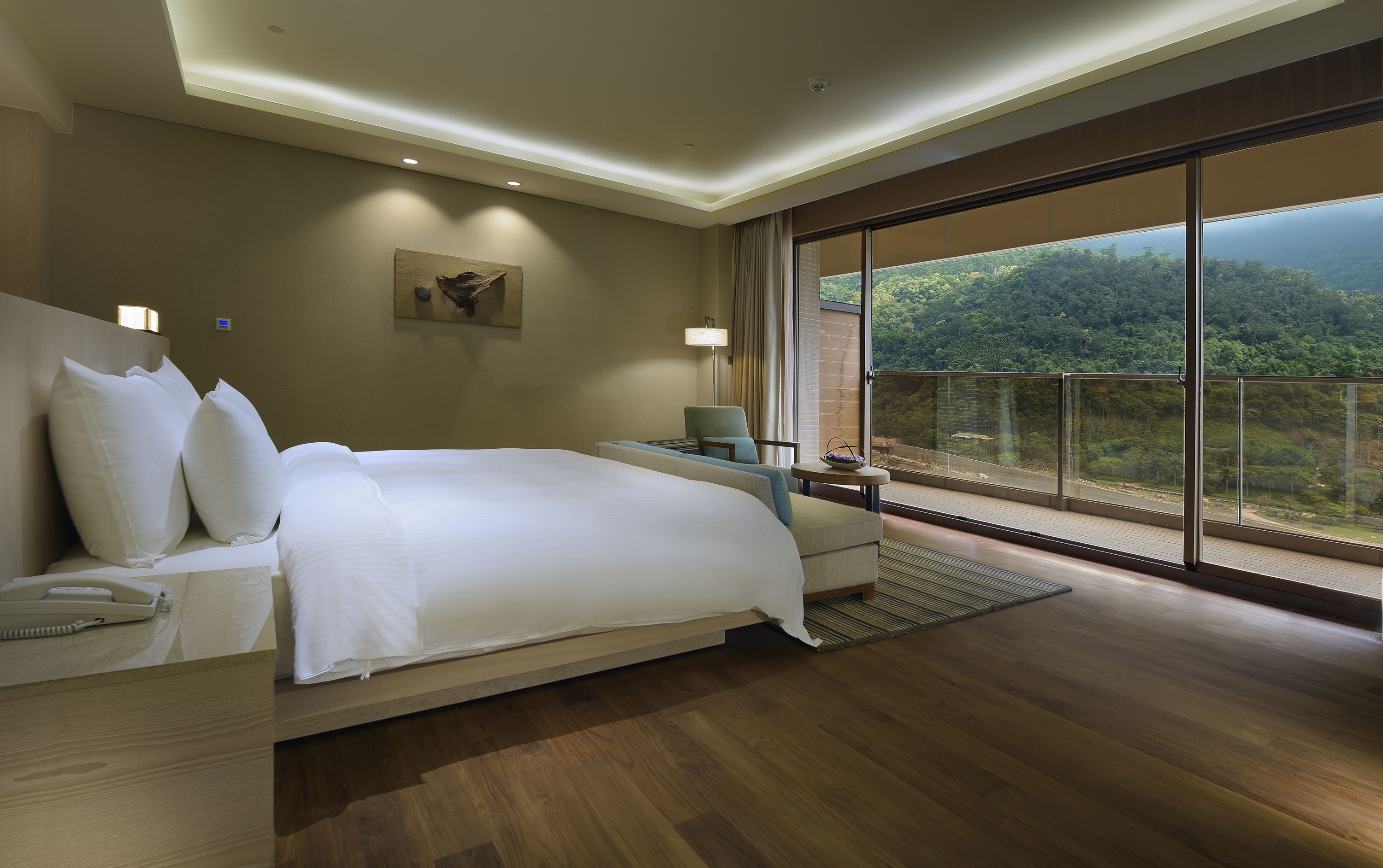 A hotel room with a large bed, a desk, a chair, and a view of a forested landscape.