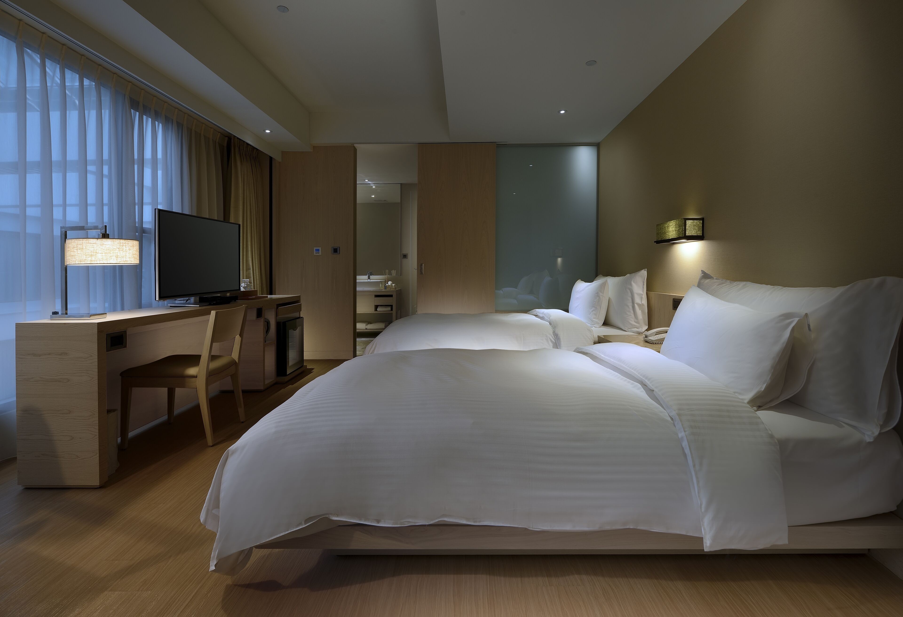 A modern hotel room with a large bed, a desk with a television, and a view of the city through large windows.