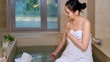 A person in a white towel sitting by a bathtub with a wooden basin containing a towel.