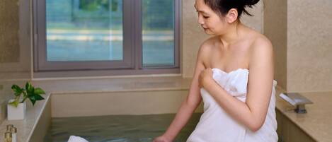Una persona con una toalla blanca sentada junto a una bañera con un lavamanos de madera que contiene una toalla.