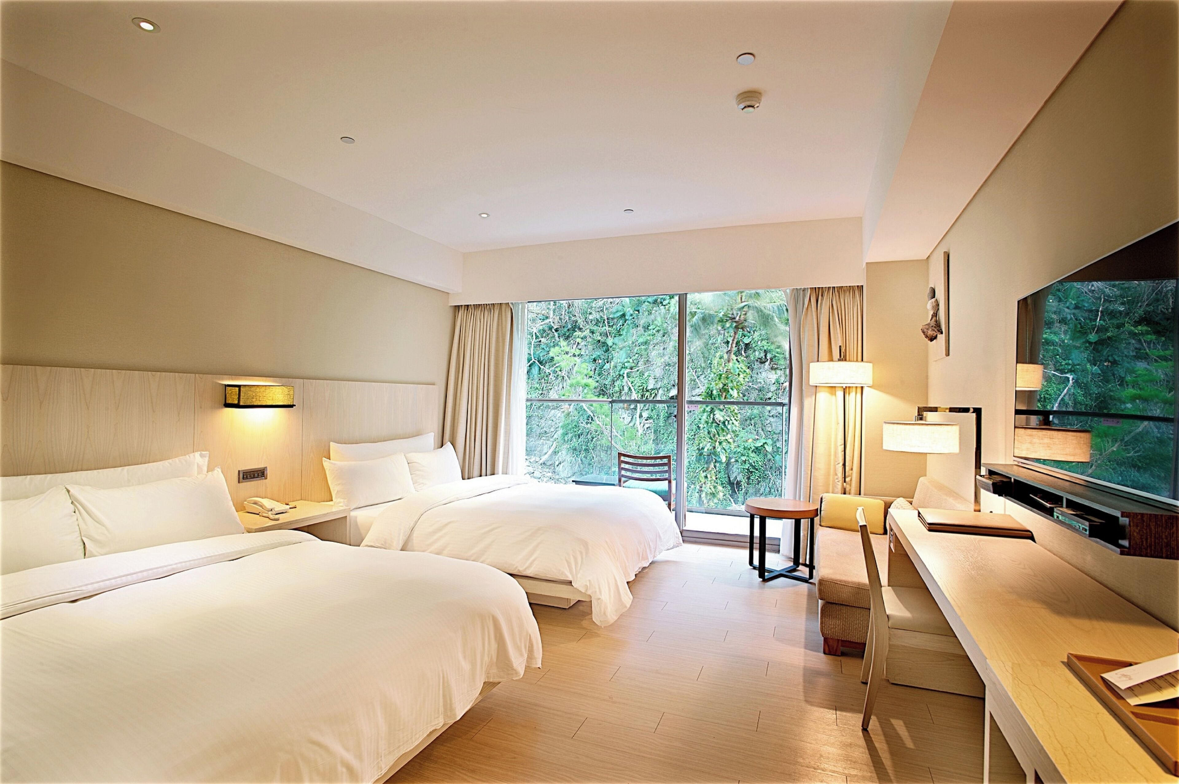 A hotel room with two beds, a desk, a TV, and a large window with a view of trees.