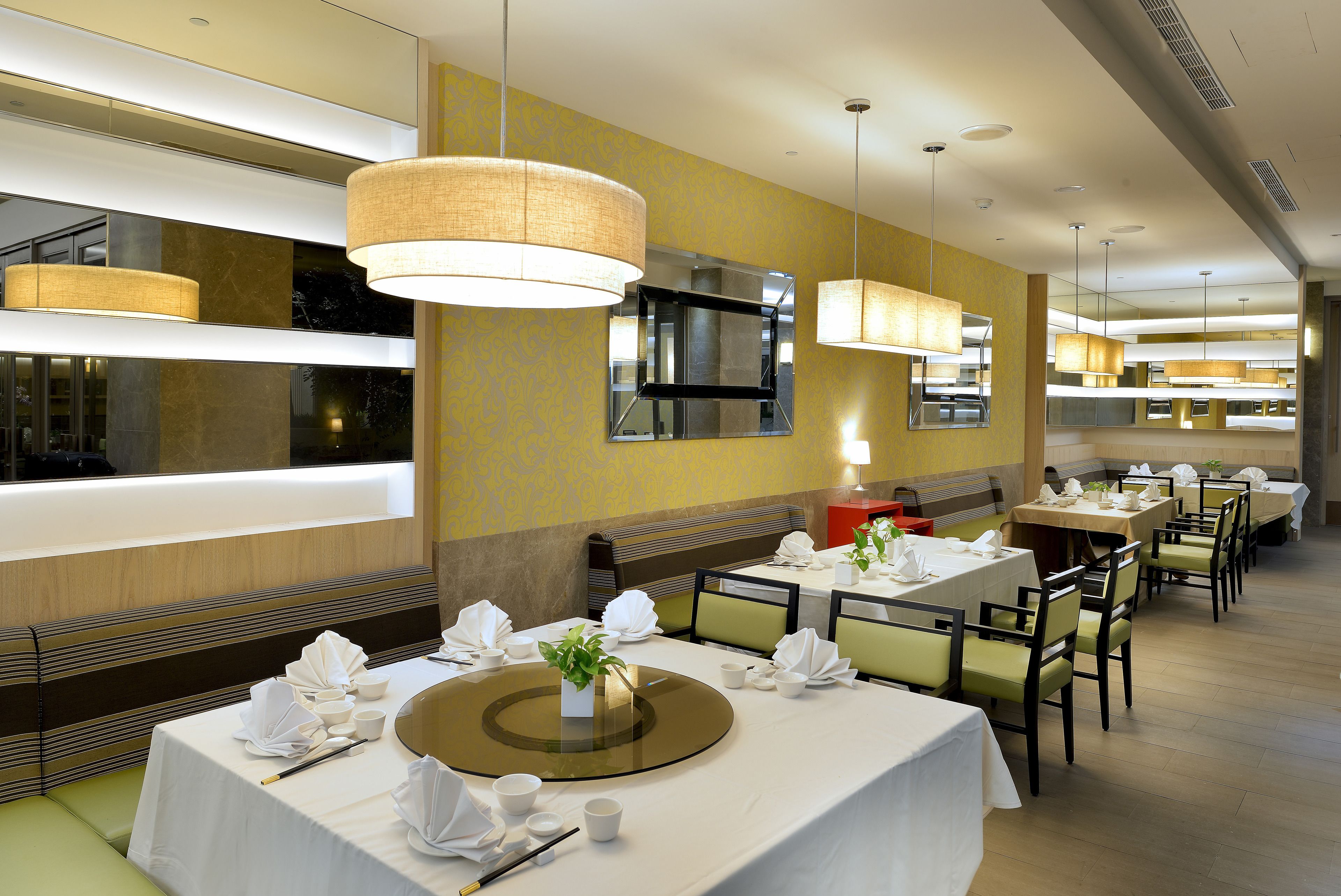 A restaurant with multiple tables set for dining, featuring white tablecloths, neatly arranged napkins, and a central decorative plant.