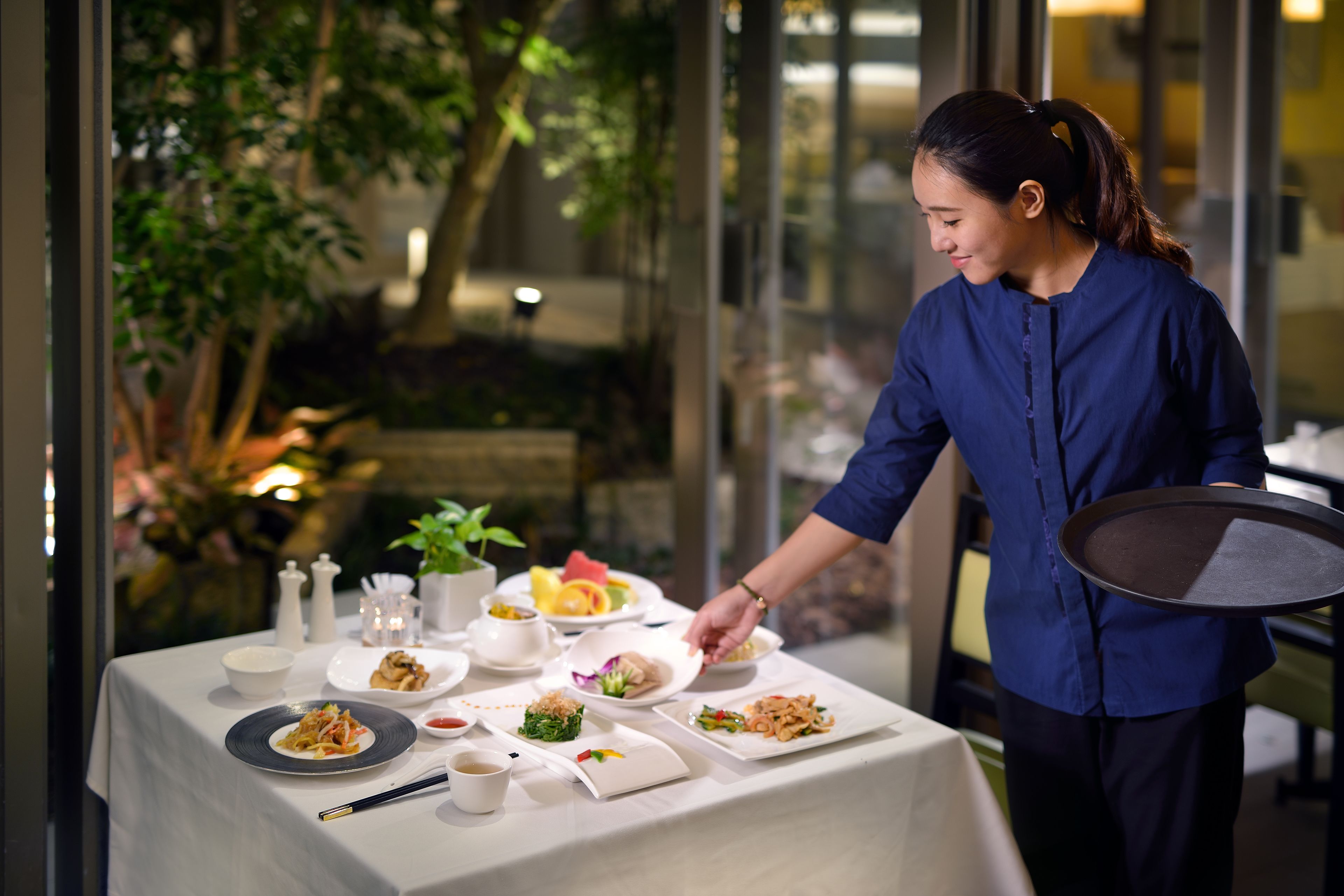 A dining table set with various dishes, a person serving food, and a view of the outdoors.