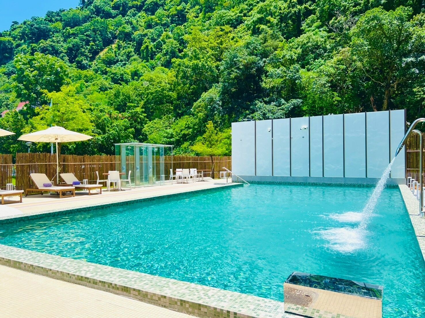 Piscina con sillas de descanso y sombrillas, rodeada por una cerca de madera y abundante vegetación.