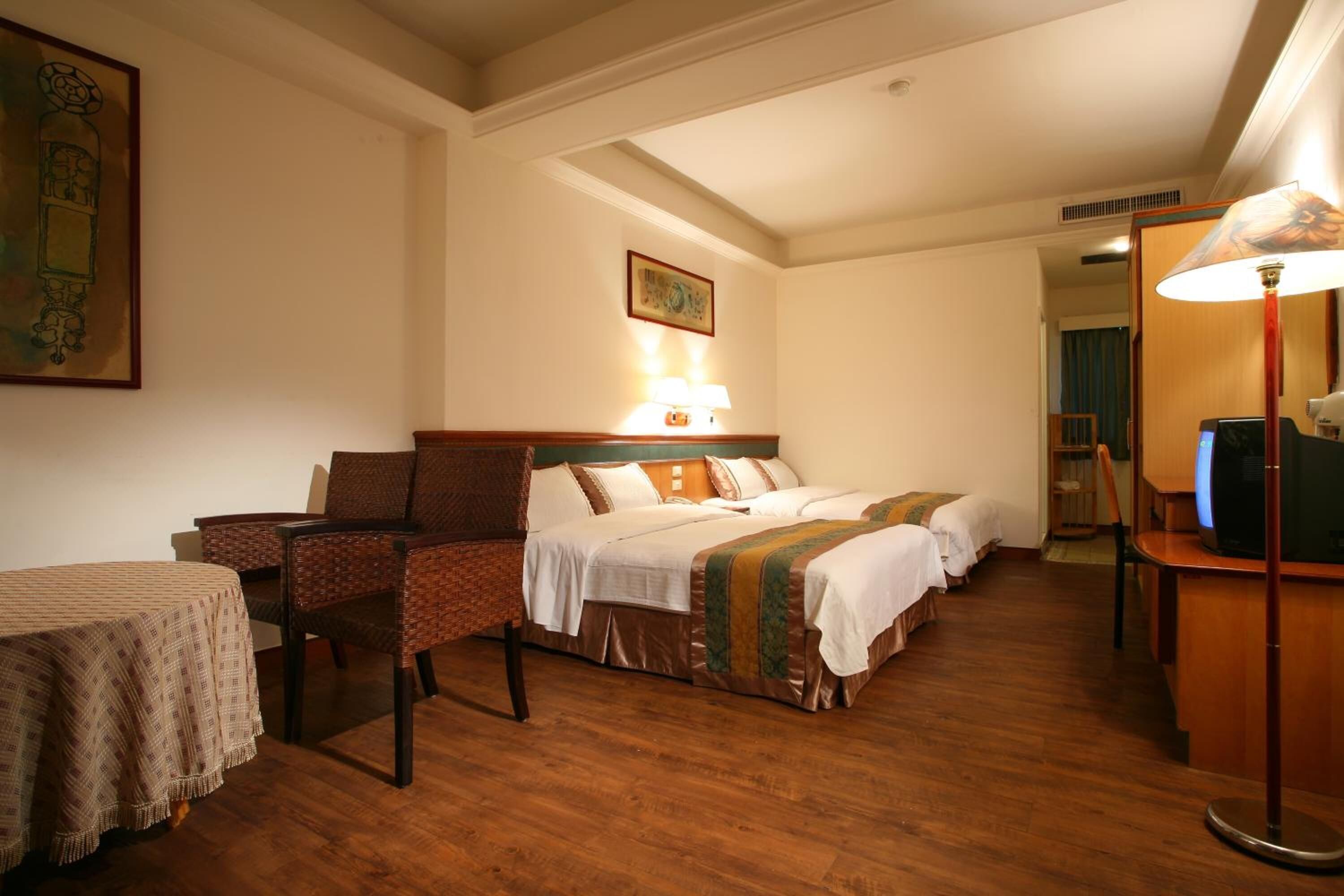 A hotel room with a wooden floor, a bed with white linens, a chair, a table with a tablecloth, and a lamp.