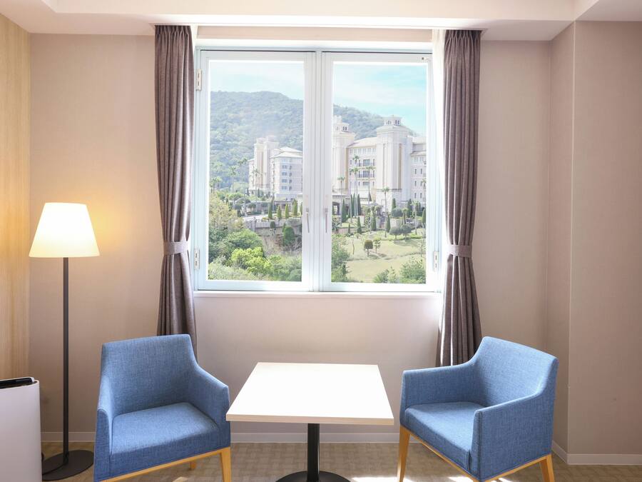 A hotel room with a large window, two blue armchairs, a small table, and a floor lamp.