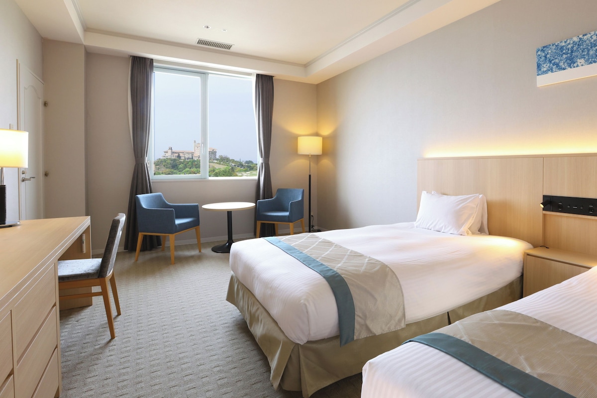 A hotel room with two beds, a desk, a chair, a table, and a large window with curtains.