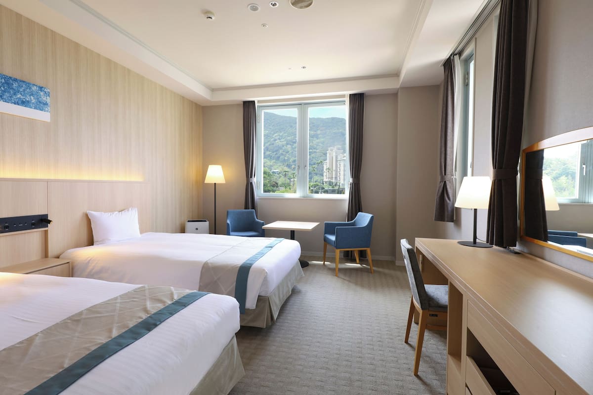 A hotel room with two beds, a desk, a chair, a lamp, and a large window with a view of greenery.