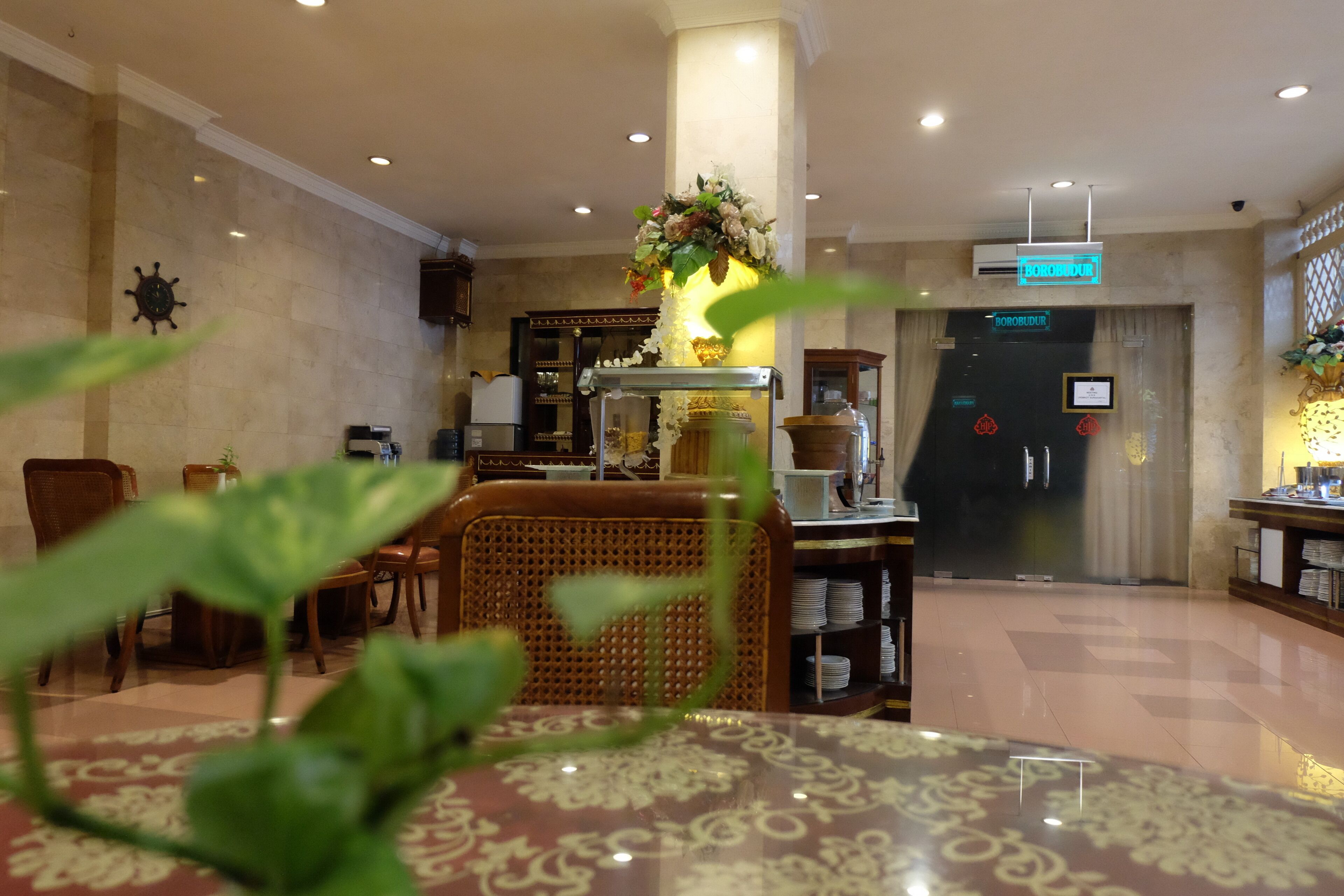 Een lobby met een receptiebalie, een koffieapparaat, rieten stoelen en een decoratieve plant.
