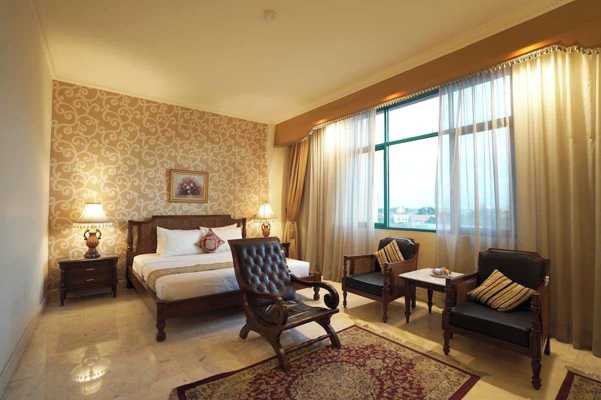 A hotel room with a bed, two chairs, a small table, a nightstand, and a window with curtains.