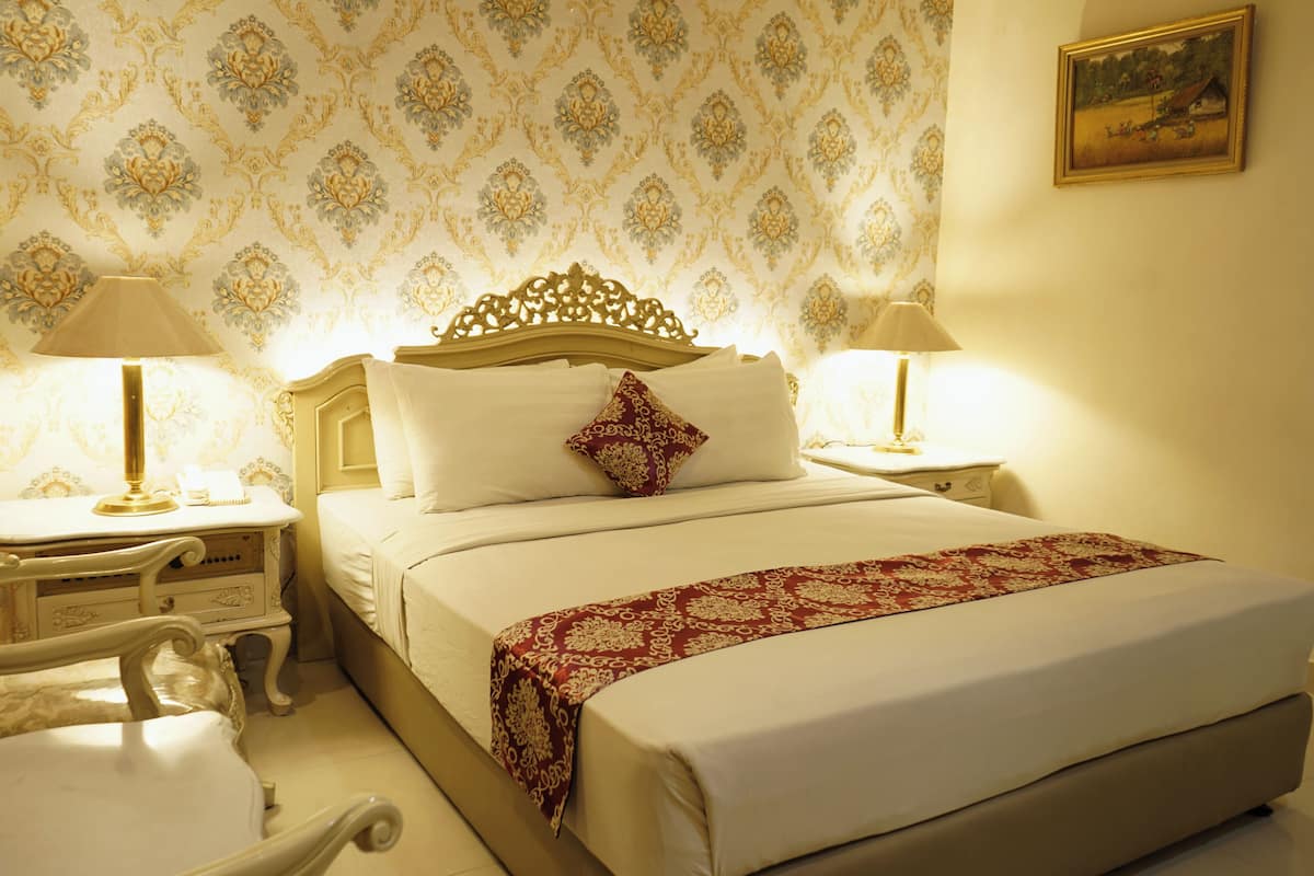 A hotel room with a large bed, ornate headboard, two bedside tables with lamps, and a patterned wallpaper.