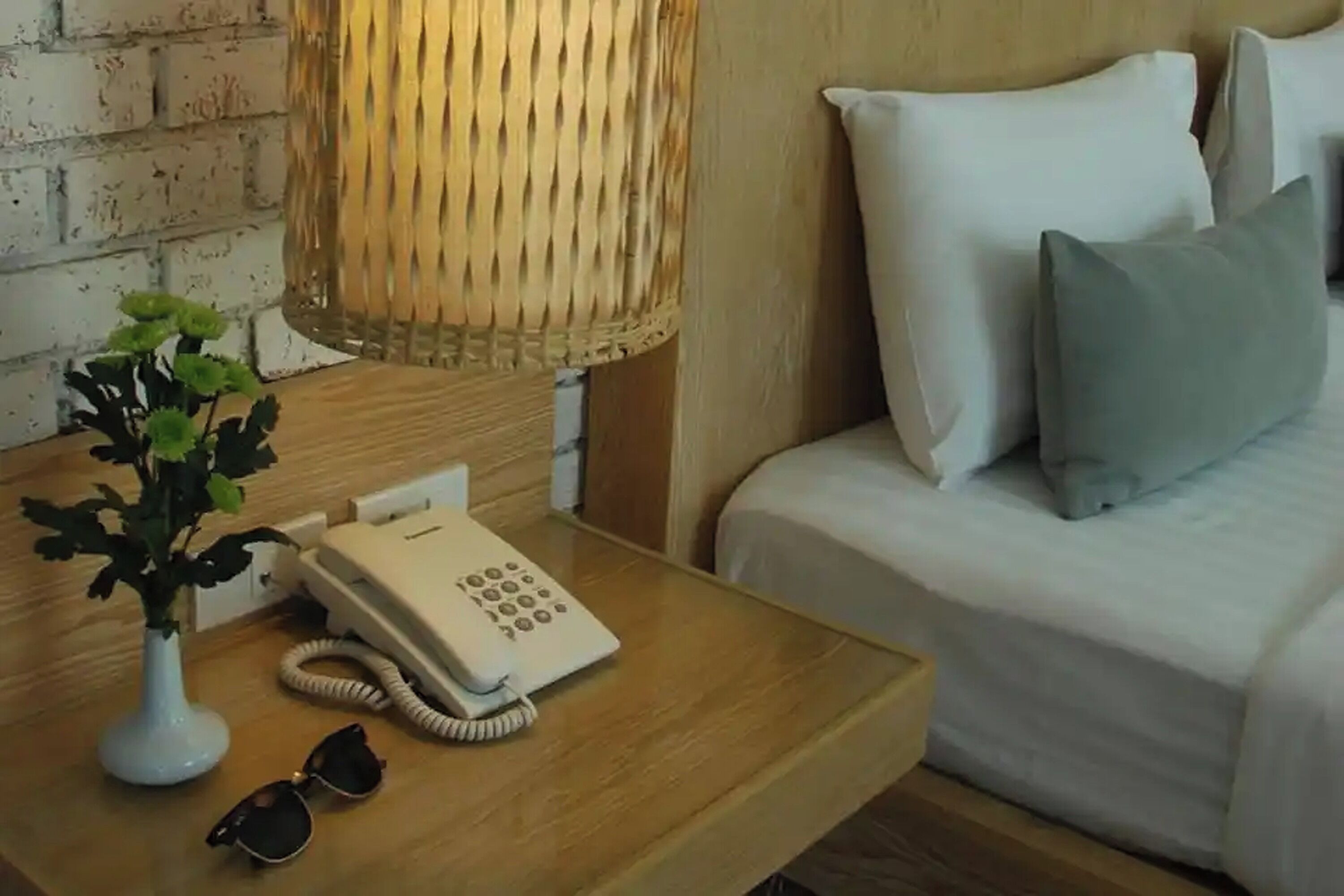 A wooden bedside table with a white telephone, a vase with green plants, and a pair of sunglasses.