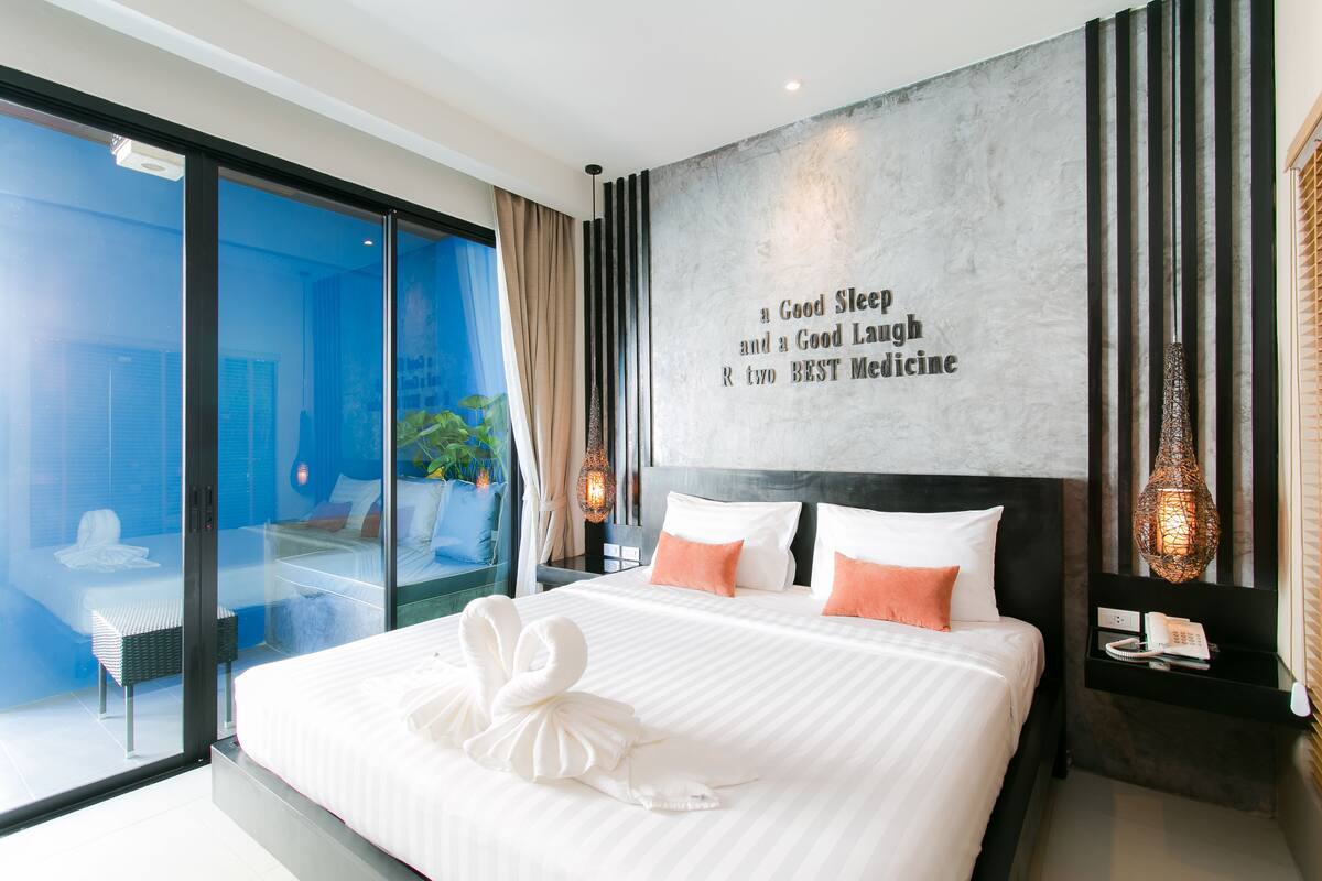 A modern hotel room with a large bed, a view through a glass door, a wall with a quote, and a bedside table with a lamp.