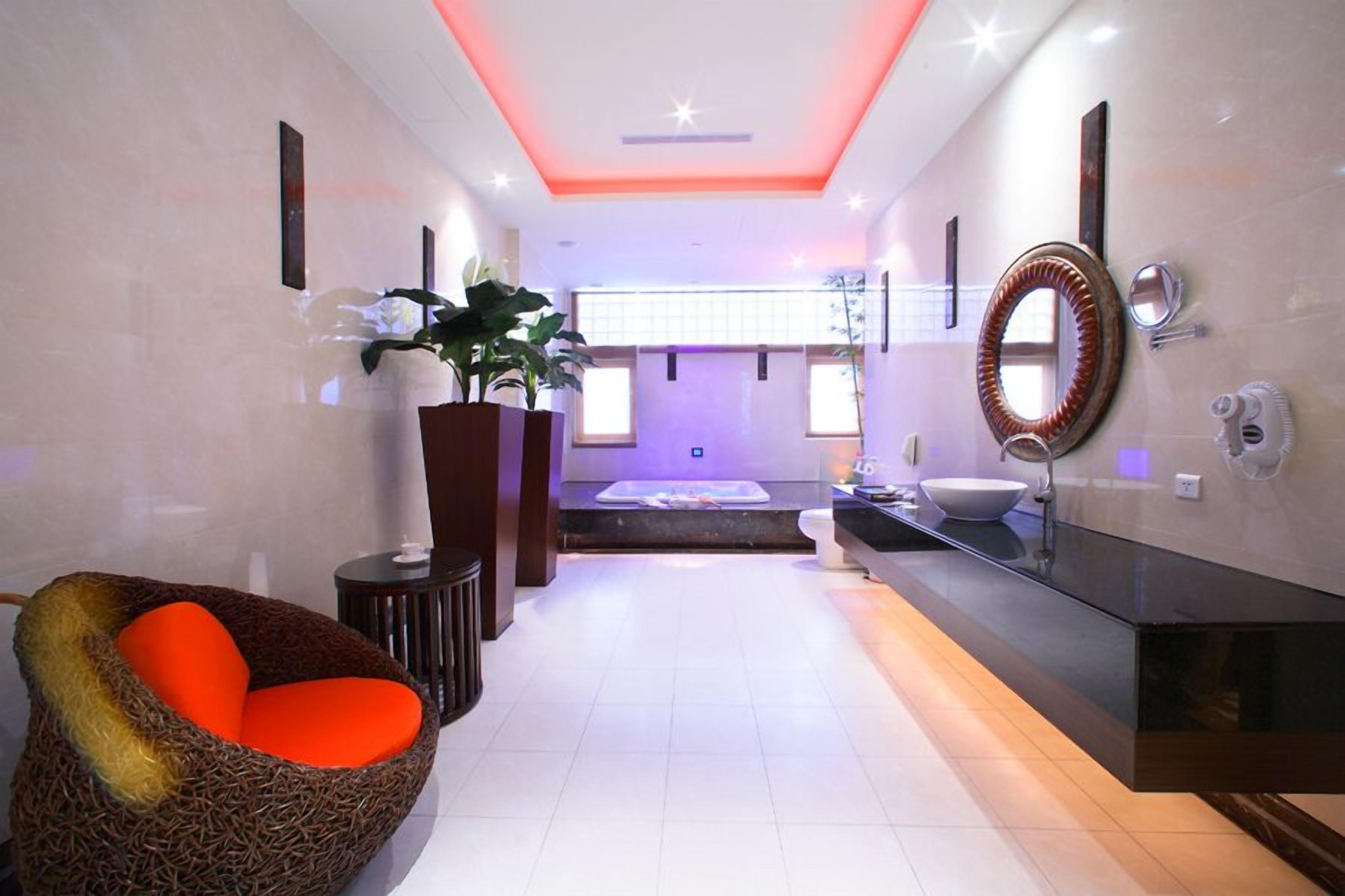A modern bathroom with a large mirror, a bathtub, and a seating area with a red cushion.