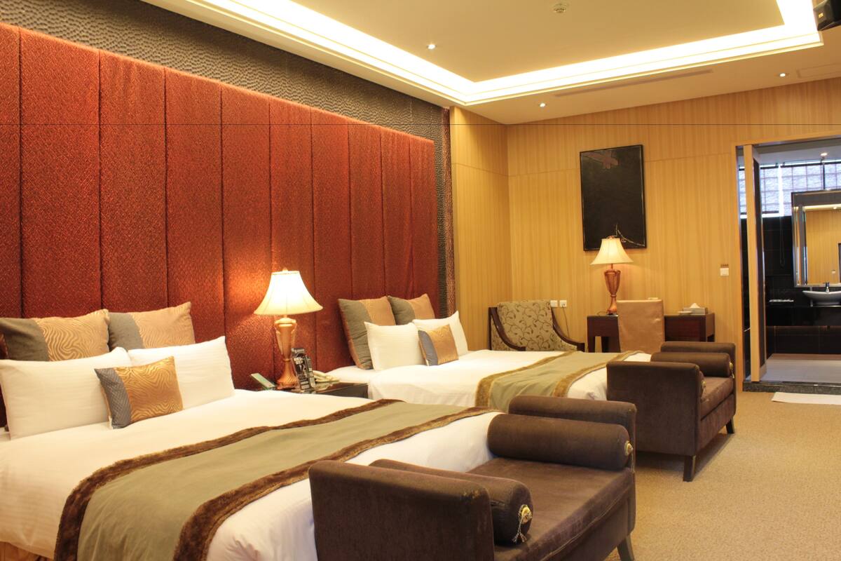 Quarto de hotel com duas camas, um sofá, uma poltrona e uma escrivaninha com abajur.