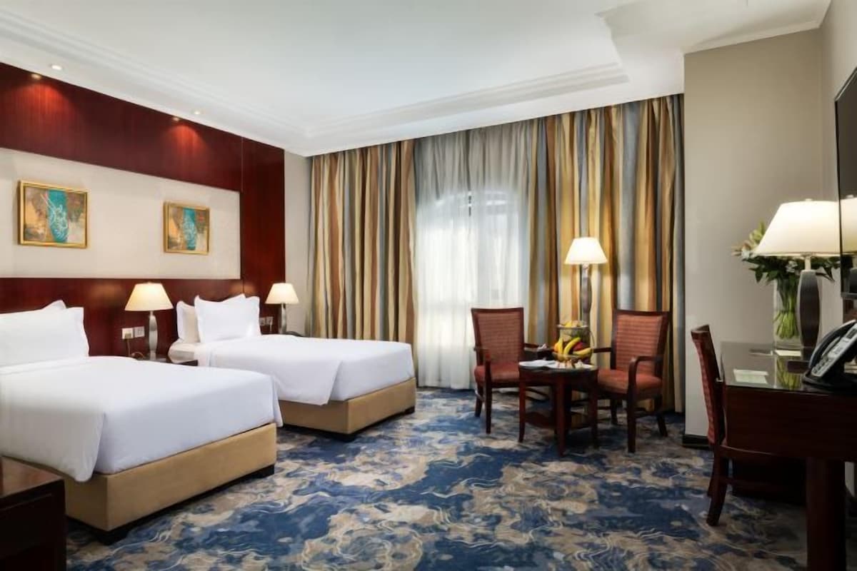 Ett hotellrum med två sängar, ett skrivbord, en stol och ett stort fönster med gardiner.