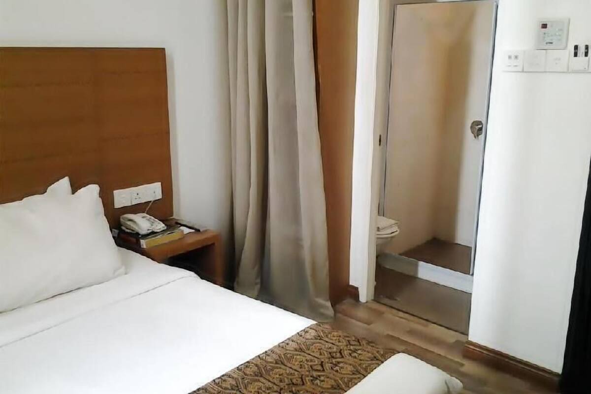 Hotellihuone, jossa on sänky, yöpöytä puhelimella, suihkutila ja seinälle asennettu termostaatti.