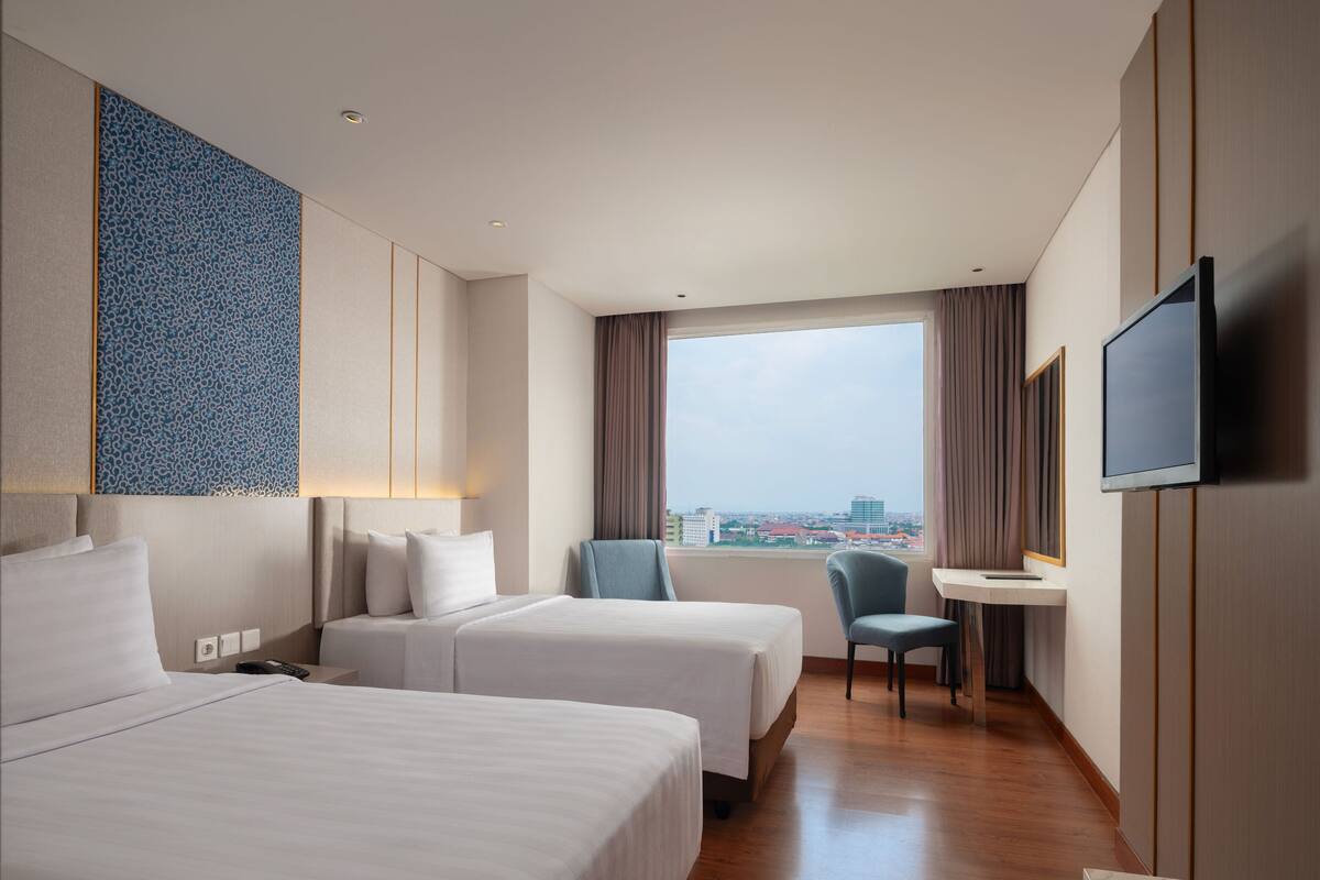Habitación de hotel con dos camas, un escritorio, una silla, televisión y un ventanal con vistas a la ciudad.