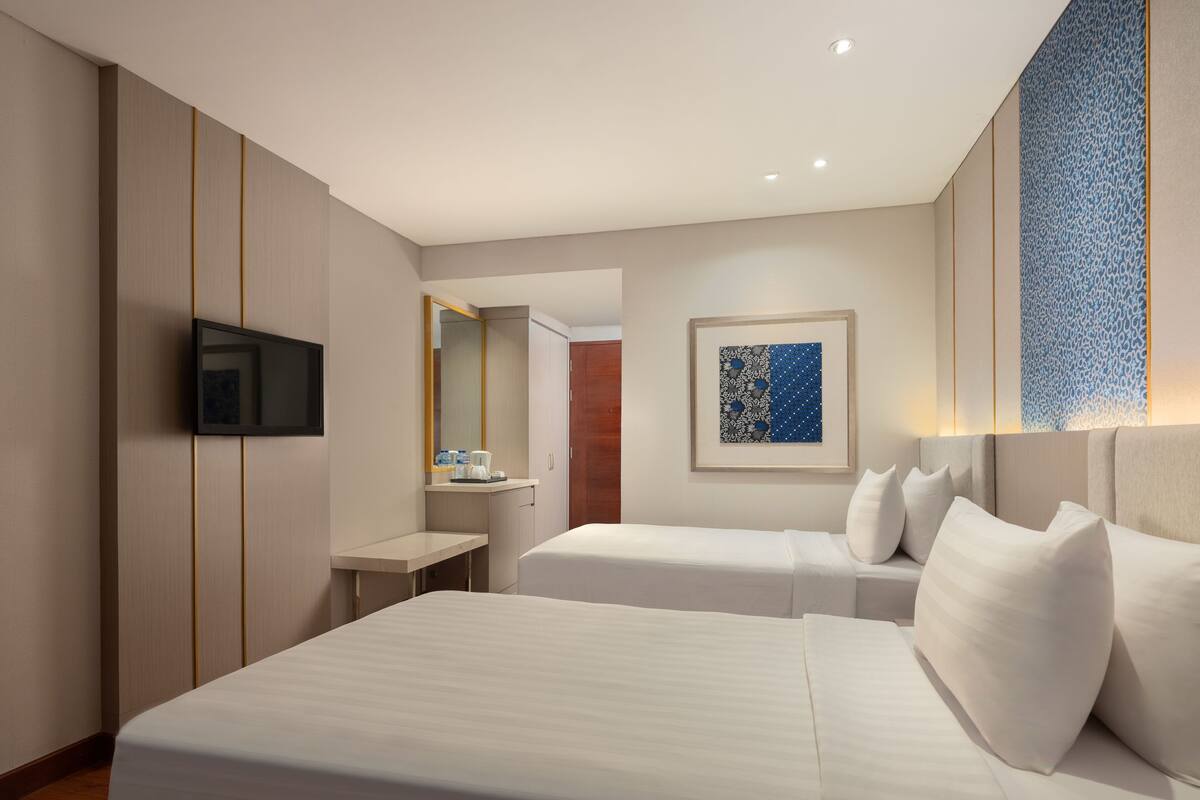 Habitación de hotel con dos camas, televisor, escritorio y baño visible a través de una puerta abierta.
