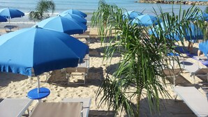 Een strand met rijen blauwe parasols en ligstoelen, een zandstrand en de zee op de achtergrond.