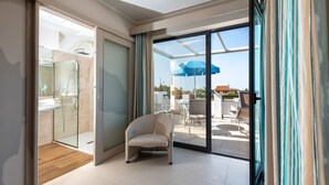 Een moderne hotelkamer met een glazen douchecabine, een zithoek met een fauteuil, en uitzicht op het buitenterras.