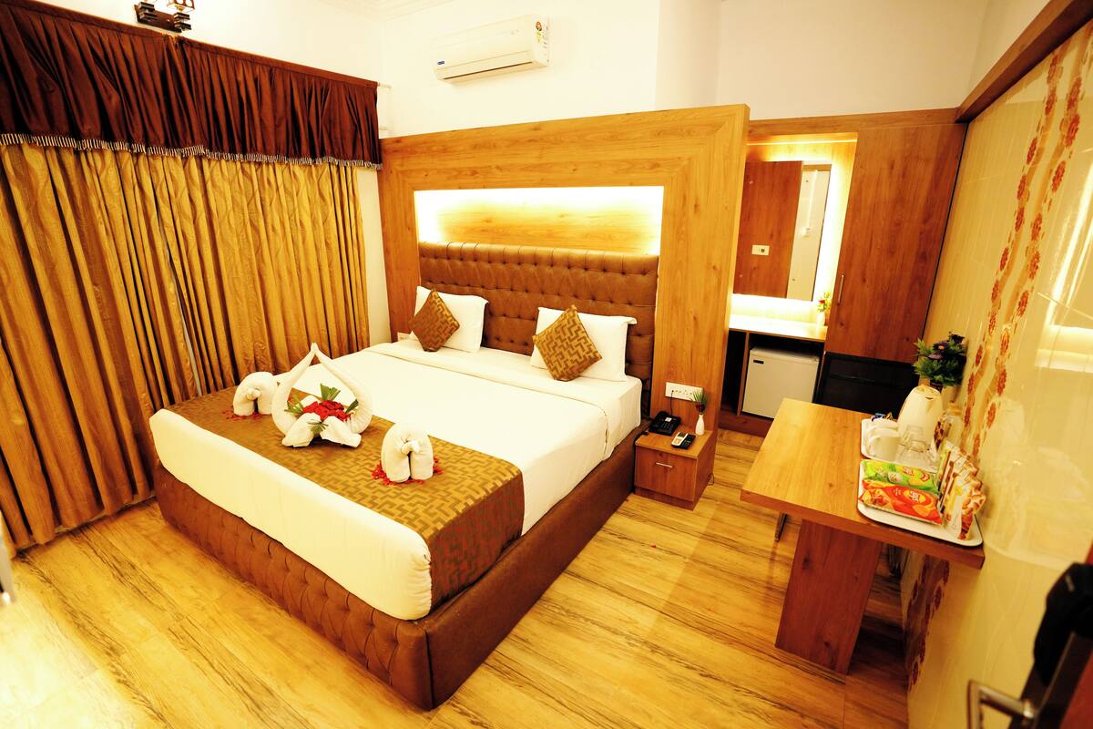 A hotel room with a large bed, a wooden headboard, a bedside table with a lamp, and a small desk with a chair.